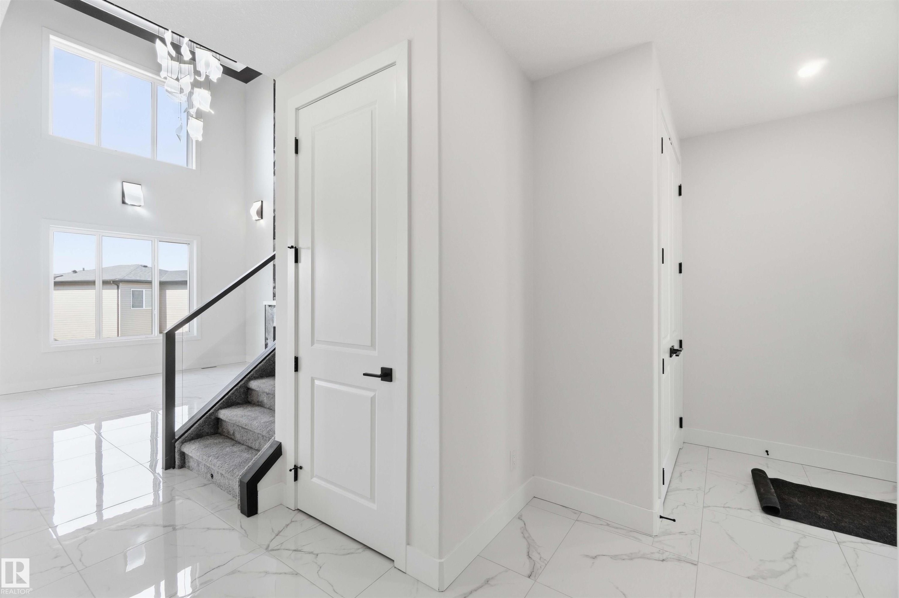 Two-story foyer featuring large windows, a contemporary chandelier, a staircase with glass railing and carpeted treads, and polished marble-look tile flooring - 415 Pine Point(E), Leduc, AB - Indoor Photo Showing Other Room