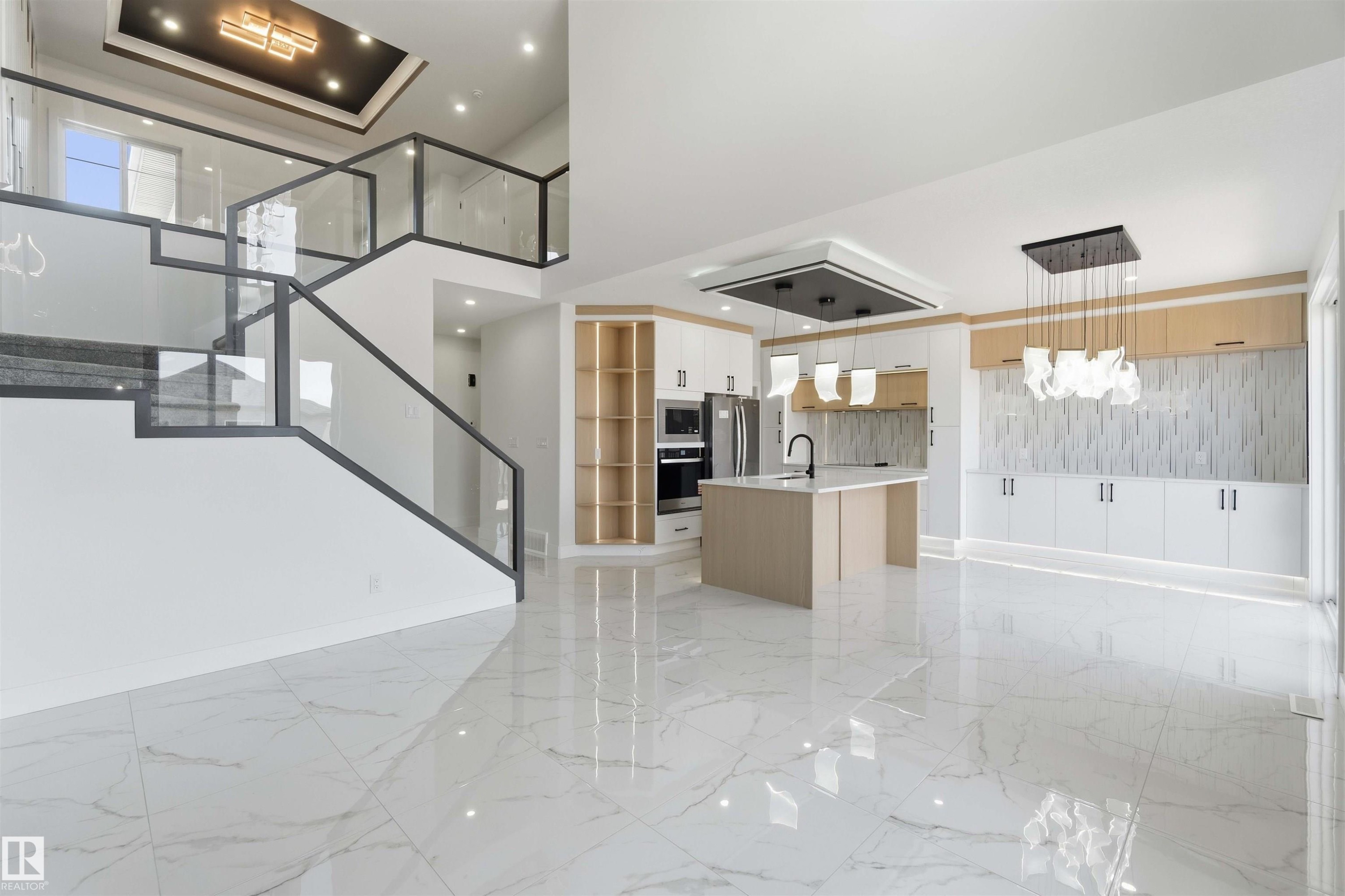 Open-concept living space featuring high-gloss marble-look floor tiles, a contemporary kitchen island with a built-in sink, integrated stainless steel appliances, and a glass-paneled staircase with dark metal railings - 415 Pine Point(E), Leduc, AB - Indoor
