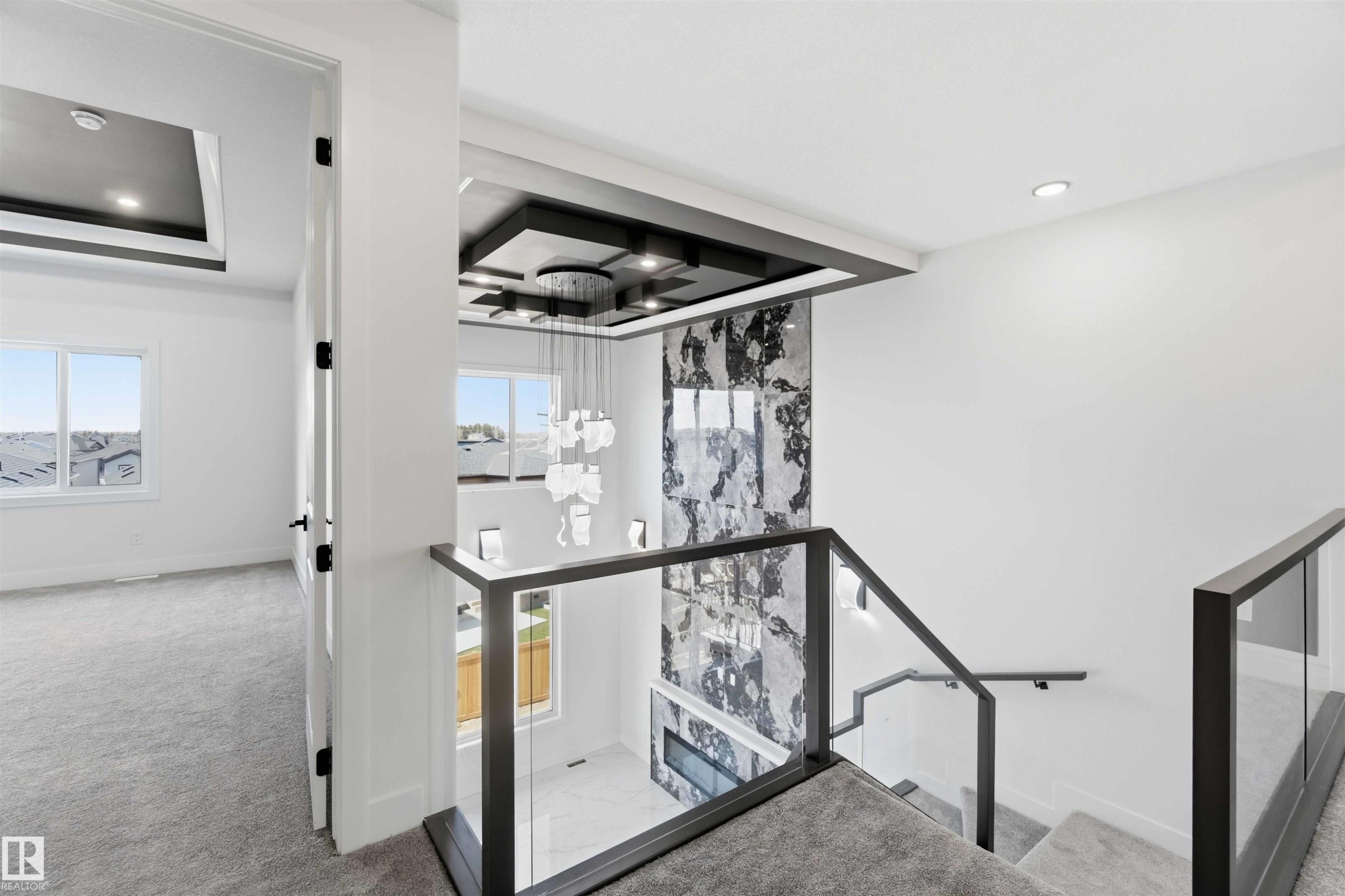 Modern staircase featuring glass panel railings with dark frames, a textured feature wall, and a multi-tiered chandelier - 415 Pine Point(E), Leduc, AB - Indoor Photo Showing Other Room