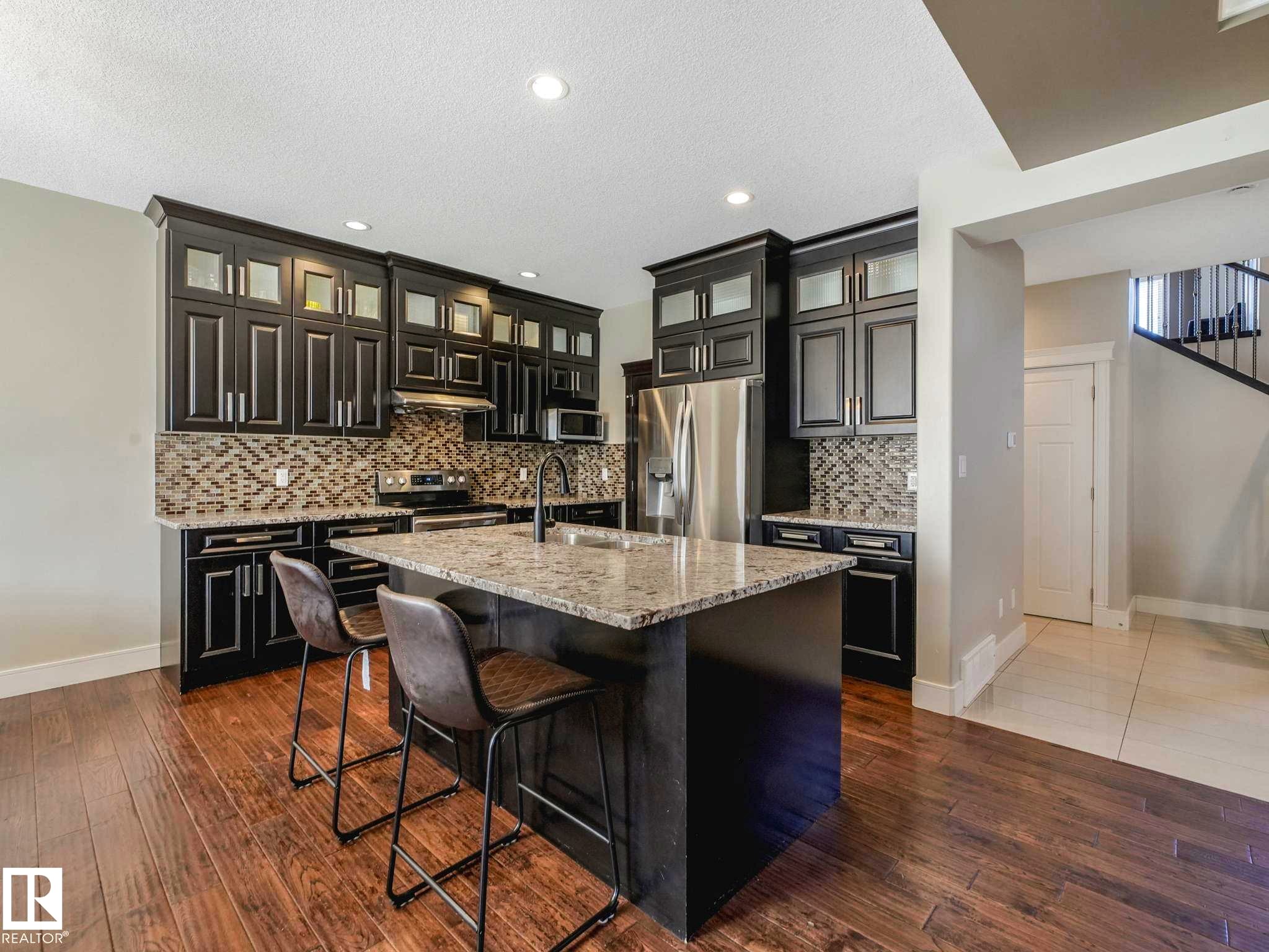 Modern kitchen featuring dark cabinetry, stainless steel appliances, a mosaic tile backsplash, and a central island with a light-toned countertop - 16251 135 Street Nw, Edmonton, AB - Indoor Photo Showing Kitchen With Stainless Steel Kitchen With Upgraded Kitchen