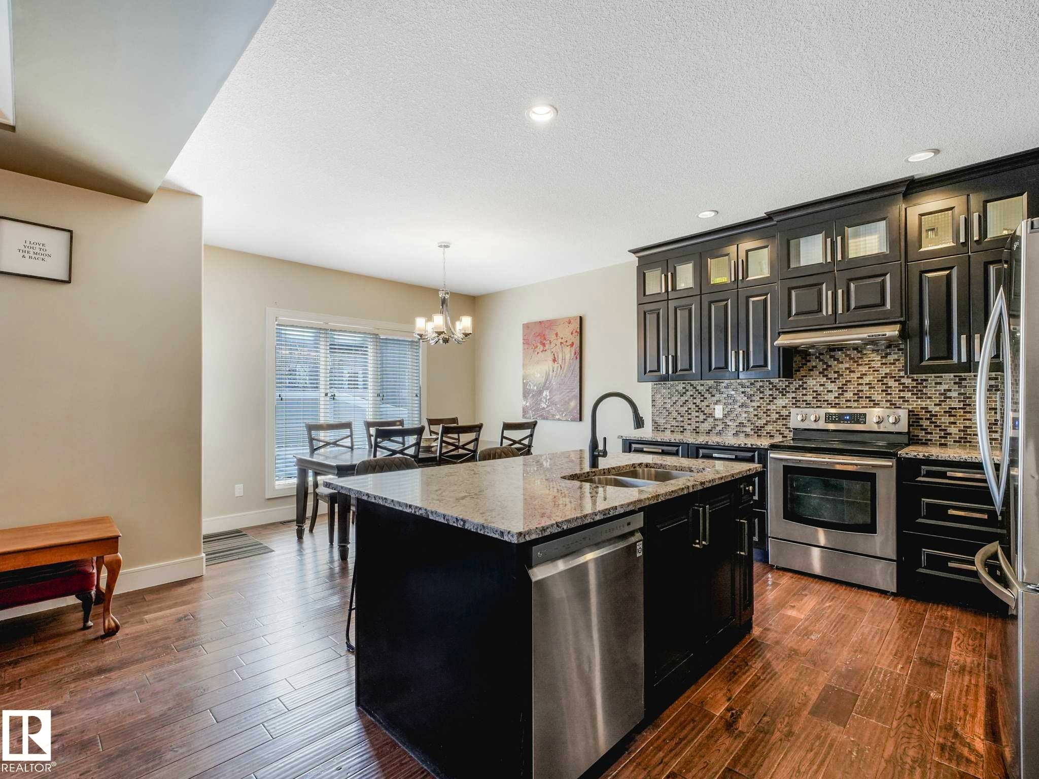 Kitchen featuring a central island with granite countertop and undermount sink, stainless steel appliances, dark cabinetry, and wood-finish flooring - 16251 135 Street Nw, Edmonton, AB - Indoor Photo Showing Kitchen With Stainless Steel Kitchen With Double Sink With Upgraded Kitchen