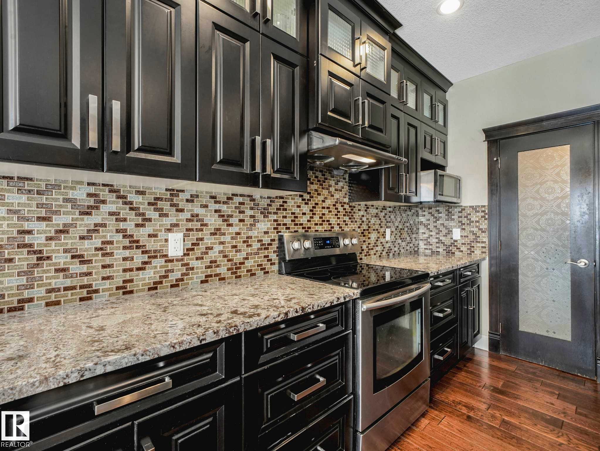 Kitchen featuring dark cabinetry, lighter countertops, a mosaic tile backsplash, stainless steel appliances, and wood-finish flooring - 16251 135 Street Nw, Edmonton, AB - Indoor Photo Showing Kitchen With Upgraded Kitchen