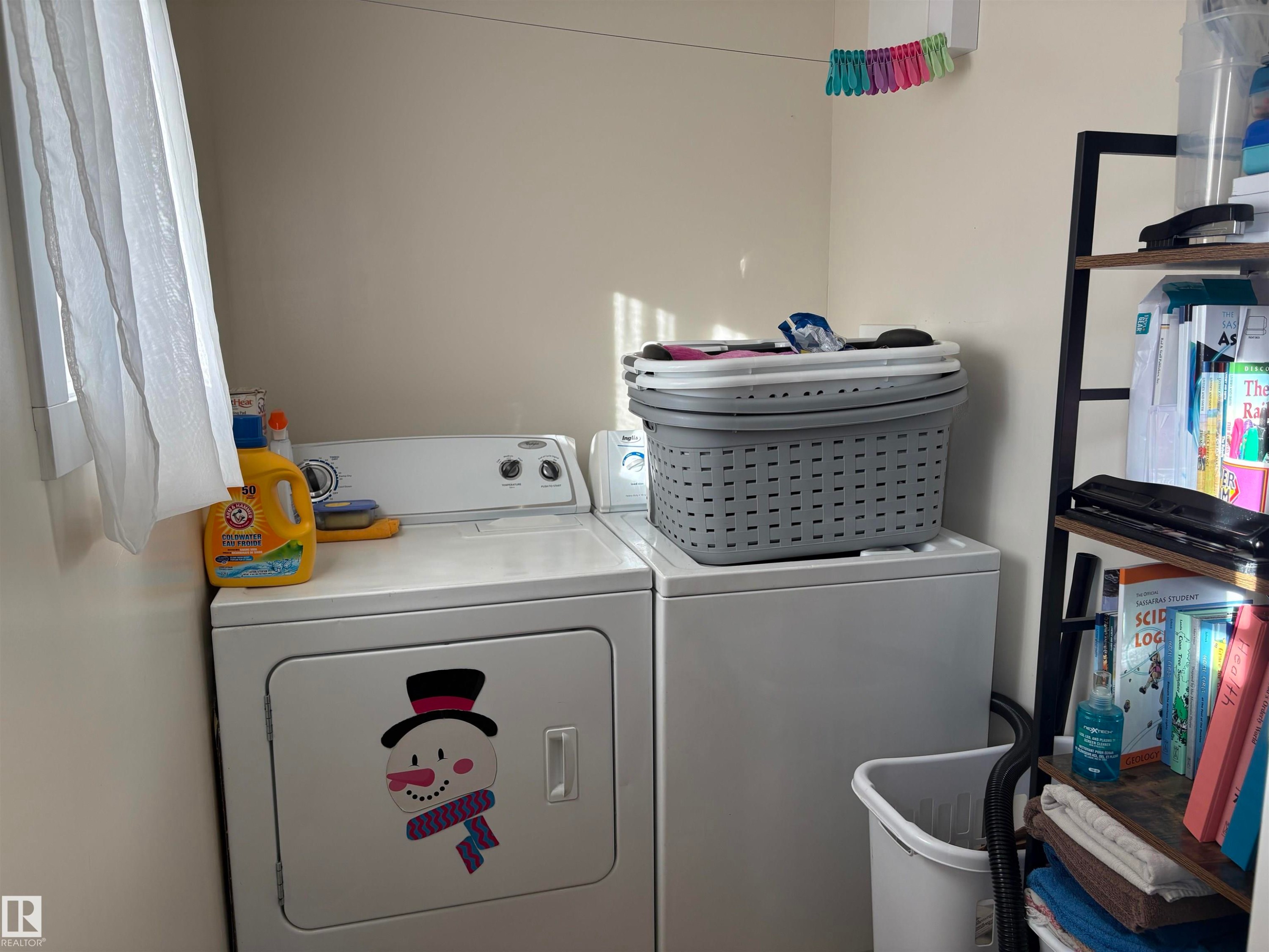 Compact laundry area featuring light-toned walls, a window with sheer curtains, and built-in shelving - 410 Main St, Alcomdale, AB - Indoor Photo Showing Laundry Room