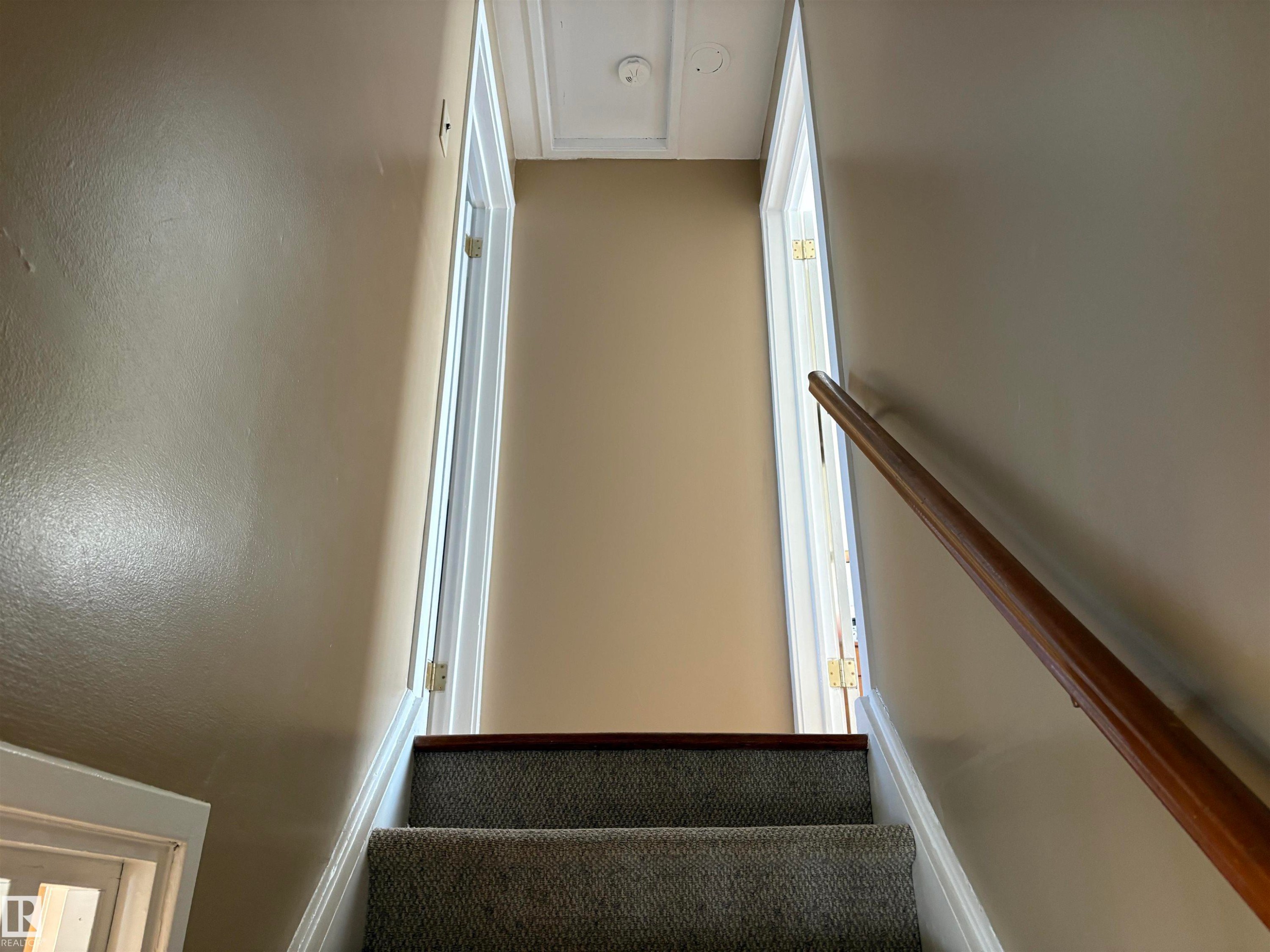 Carpeted staircase with a wood handrail, light beige wall paint, white trim, and a ceiling access panel - 410 Main St, Alcomdale, AB - Indoor Photo Showing Other Room