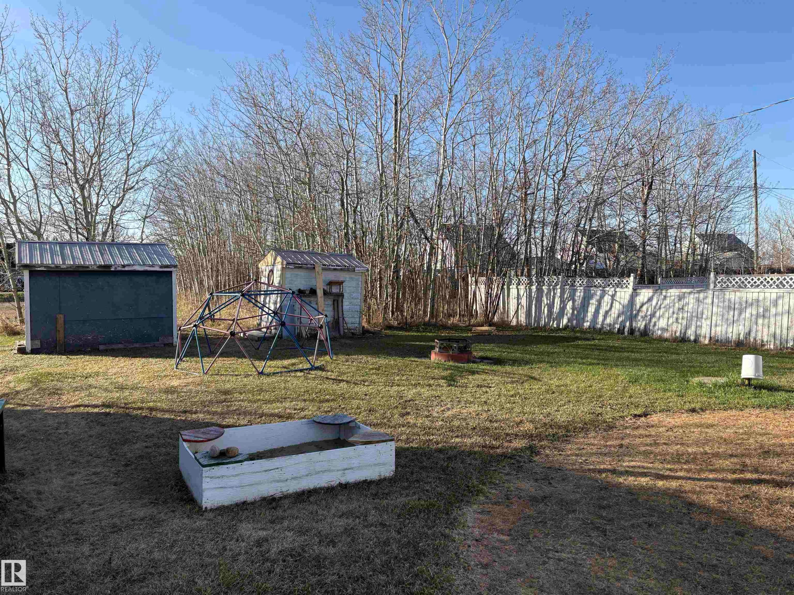 Expansive outdoor area featuring two storage sheds with metal roofs, a white privacy fence, a raised garden bed, and a circular fire pit - 410 Main St, Alcomdale, AB - Outdoor