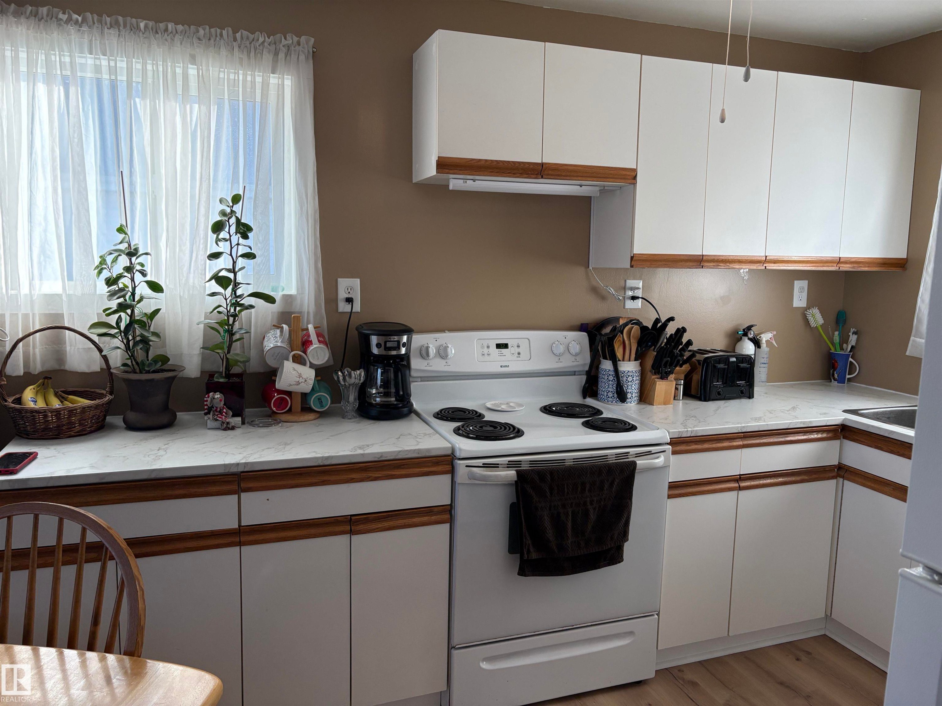 Kitchen featuring white cabinetry with wood-finish trim, white appliances, and a window with sheer curtains - 410 Main St, Alcomdale, AB - Indoor Photo Showing Kitchen