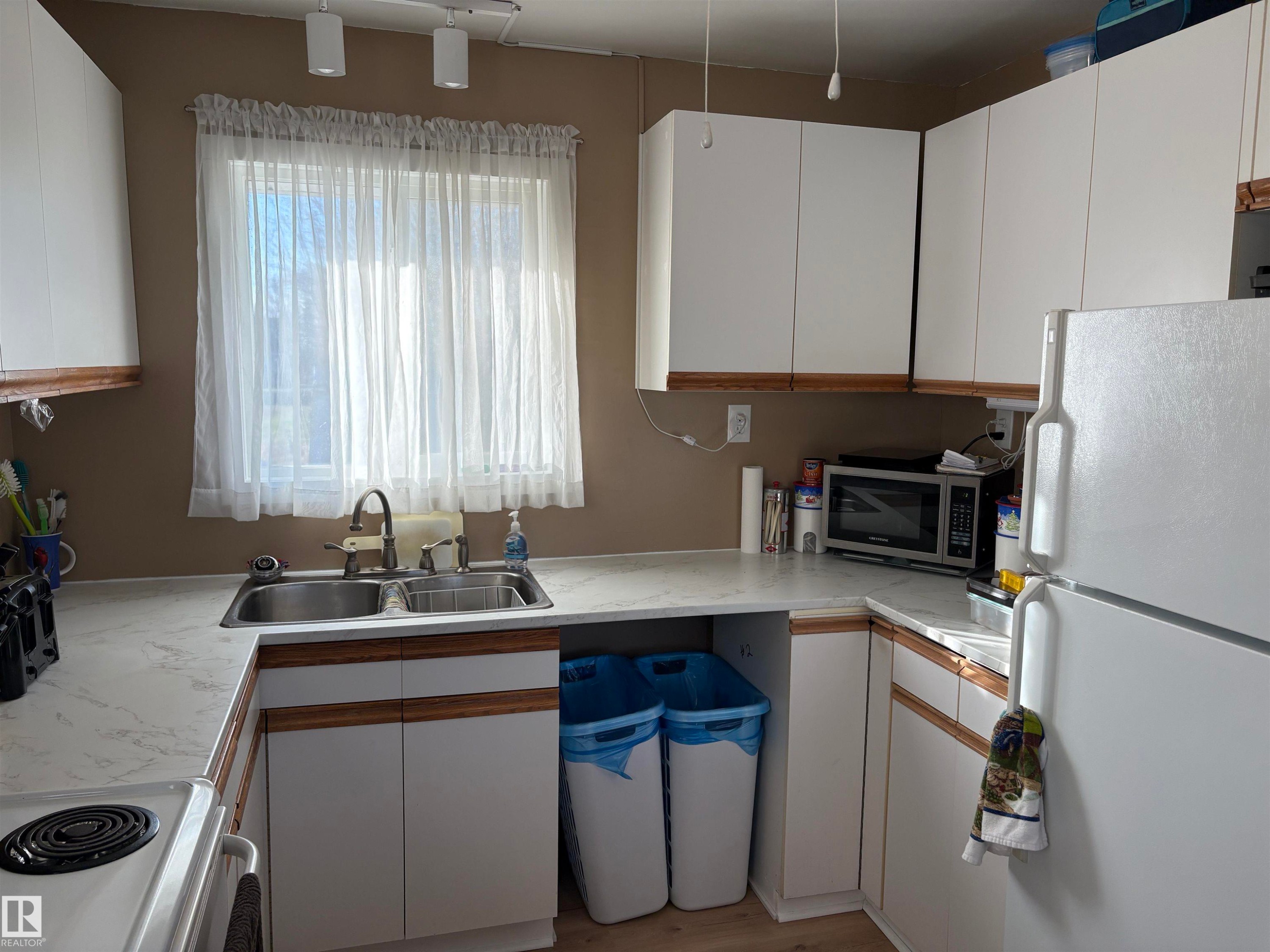 Functional kitchen featuring white cabinetry with wood-finish trim, a double basin stainless steel sink, and light-toned countertops - 410 Main St, Alcomdale, AB - Indoor Photo Showing Kitchen With Double Sink