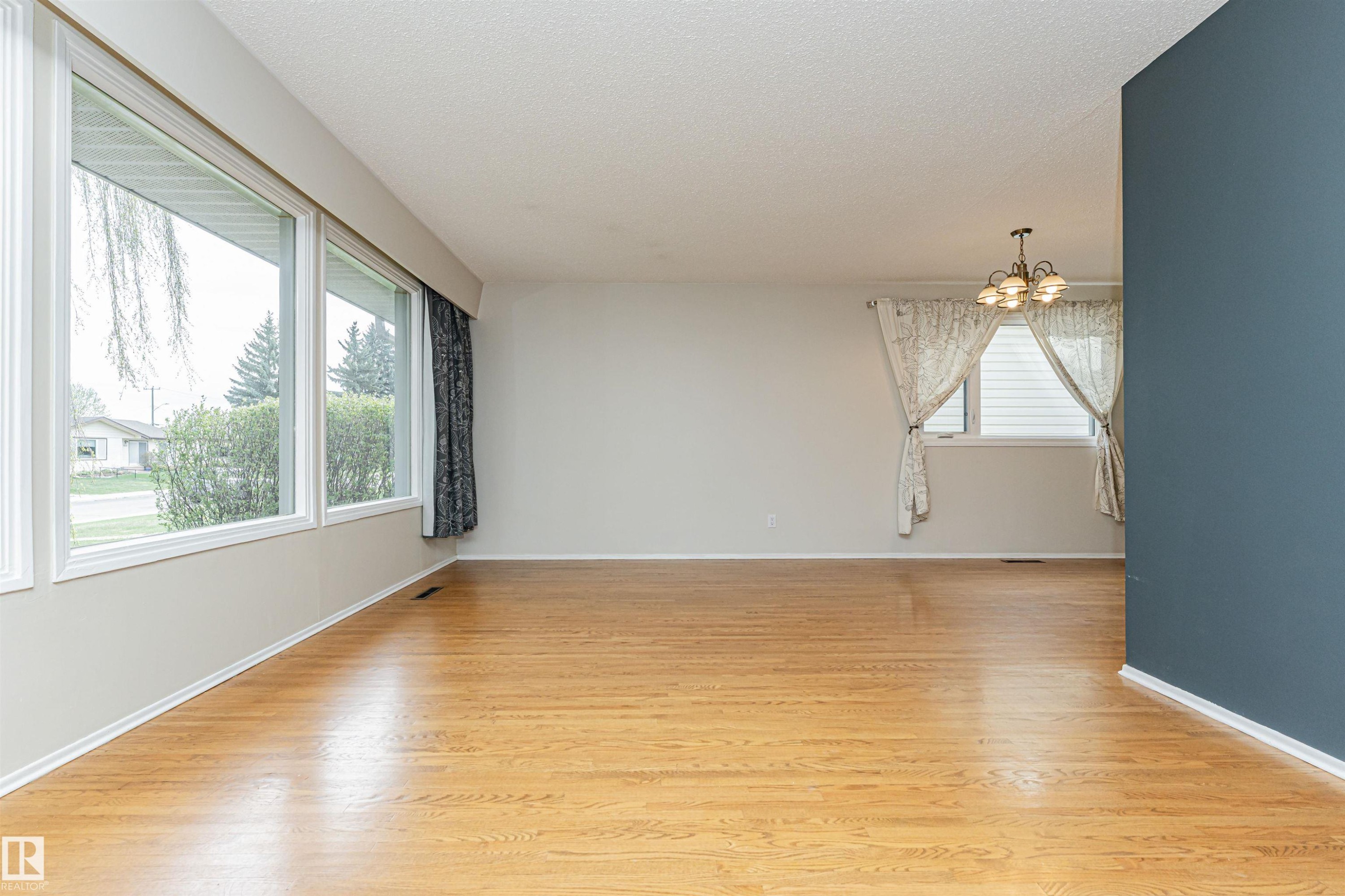 Spacious room featuring wood-finish flooring, large picture windows, a ceiling-mounted light fixture, and a partial accent wall - 11423 50 Avenue, Edmonton, AB - Indoor Photo Showing Other Room