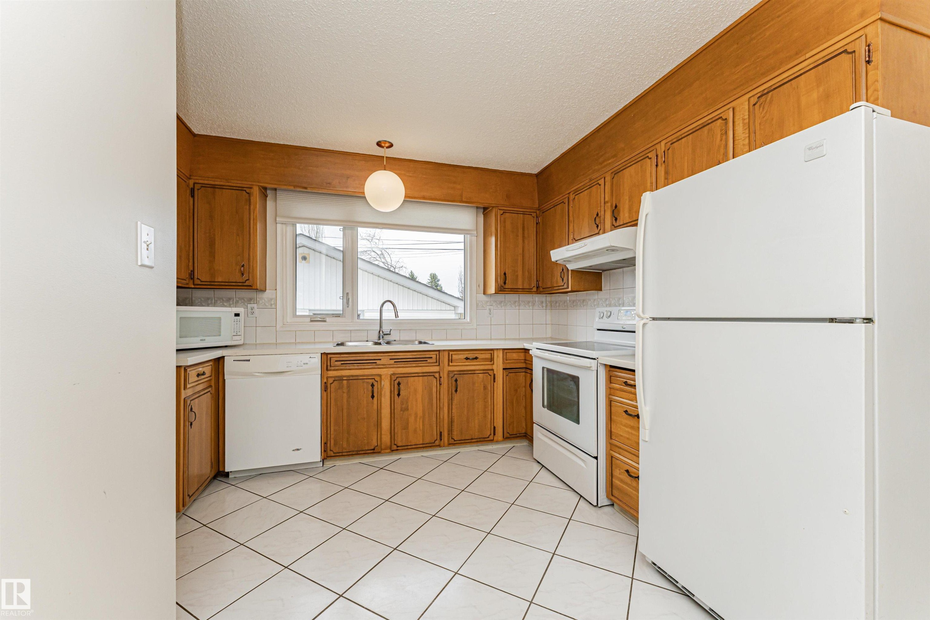 Kitchen featuring wood cabinetry, white tile flooring, white appliances, a large window above the sink, and a globe pendant light fixture - 11423 50 Avenue, Edmonton, AB - Indoor Photo Showing Kitchen With Double Sink