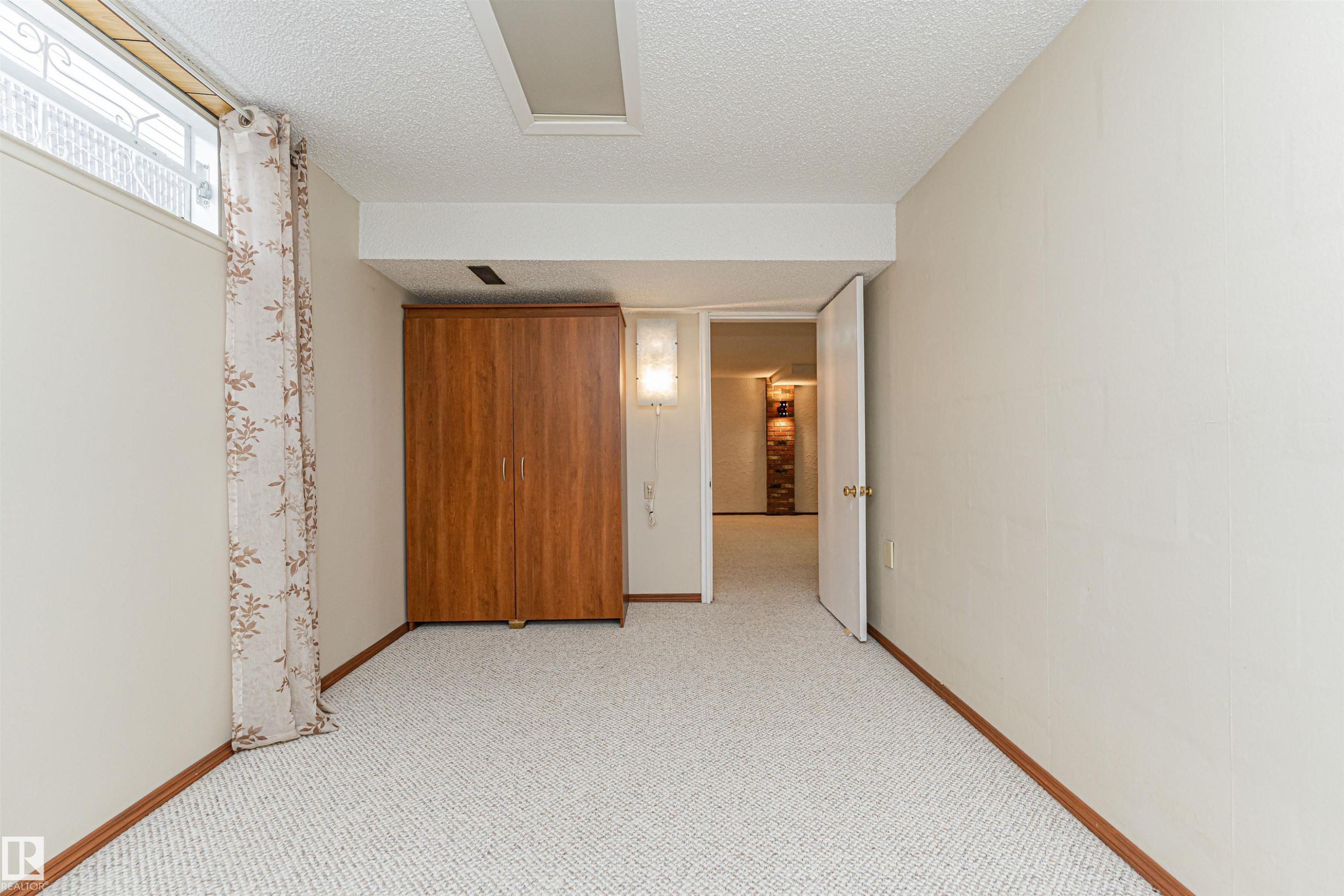 Carpeted room featuring a drop ceiling, a wood-finish armoire, a wall-mounted light fixture, a high horizontal window, and a doorway opening to an interior space - 11423 50 Avenue, Edmonton, AB - Indoor Photo Showing Other Room