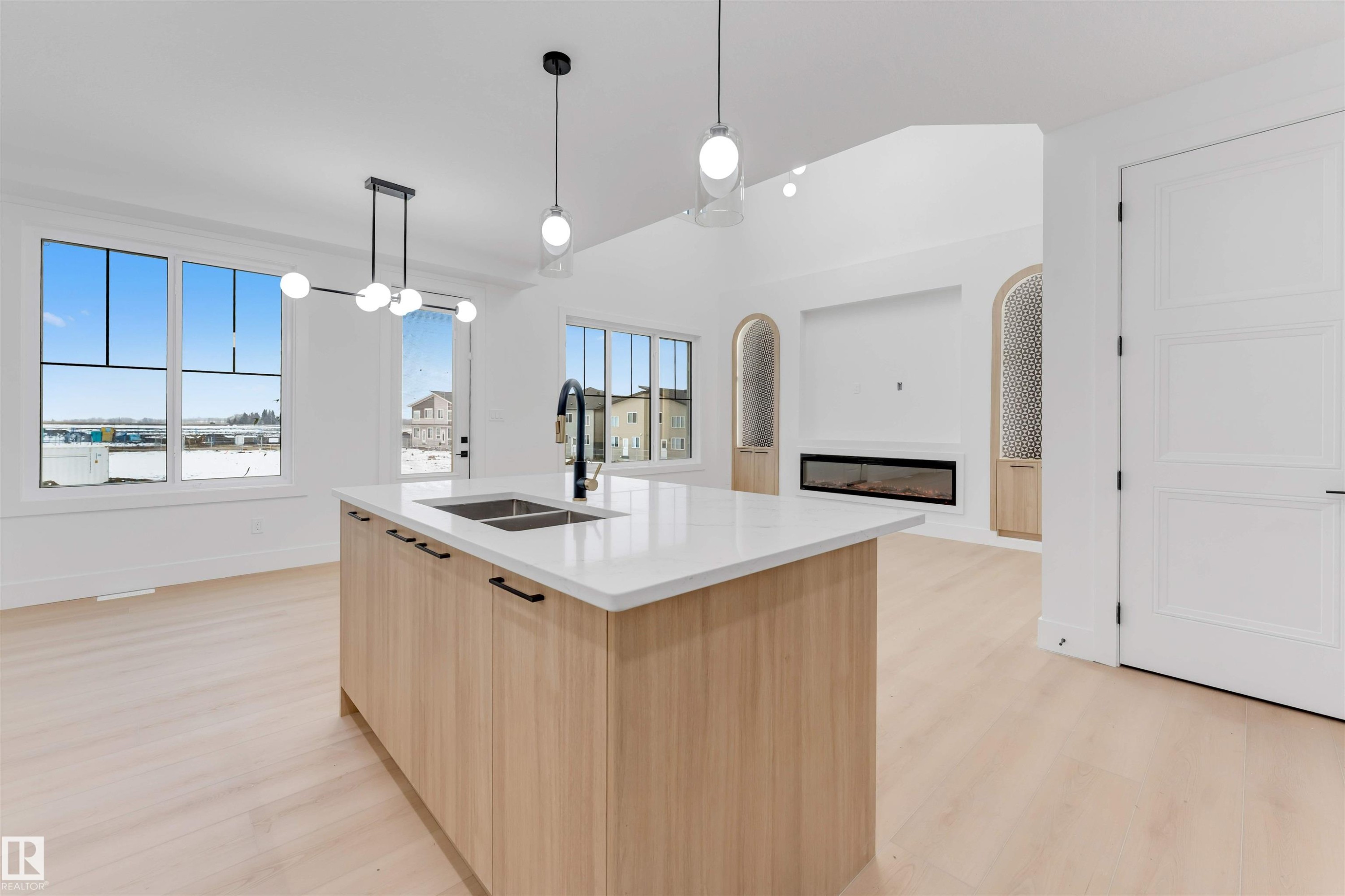 This open-concept living space features light wood flooring, a central kitchen island with a double sink and white countertop, and a fireplace with arched built-in features - 4004 35 Street, Beaumont, AB - Indoor Photo Showing Kitchen With Double Sink