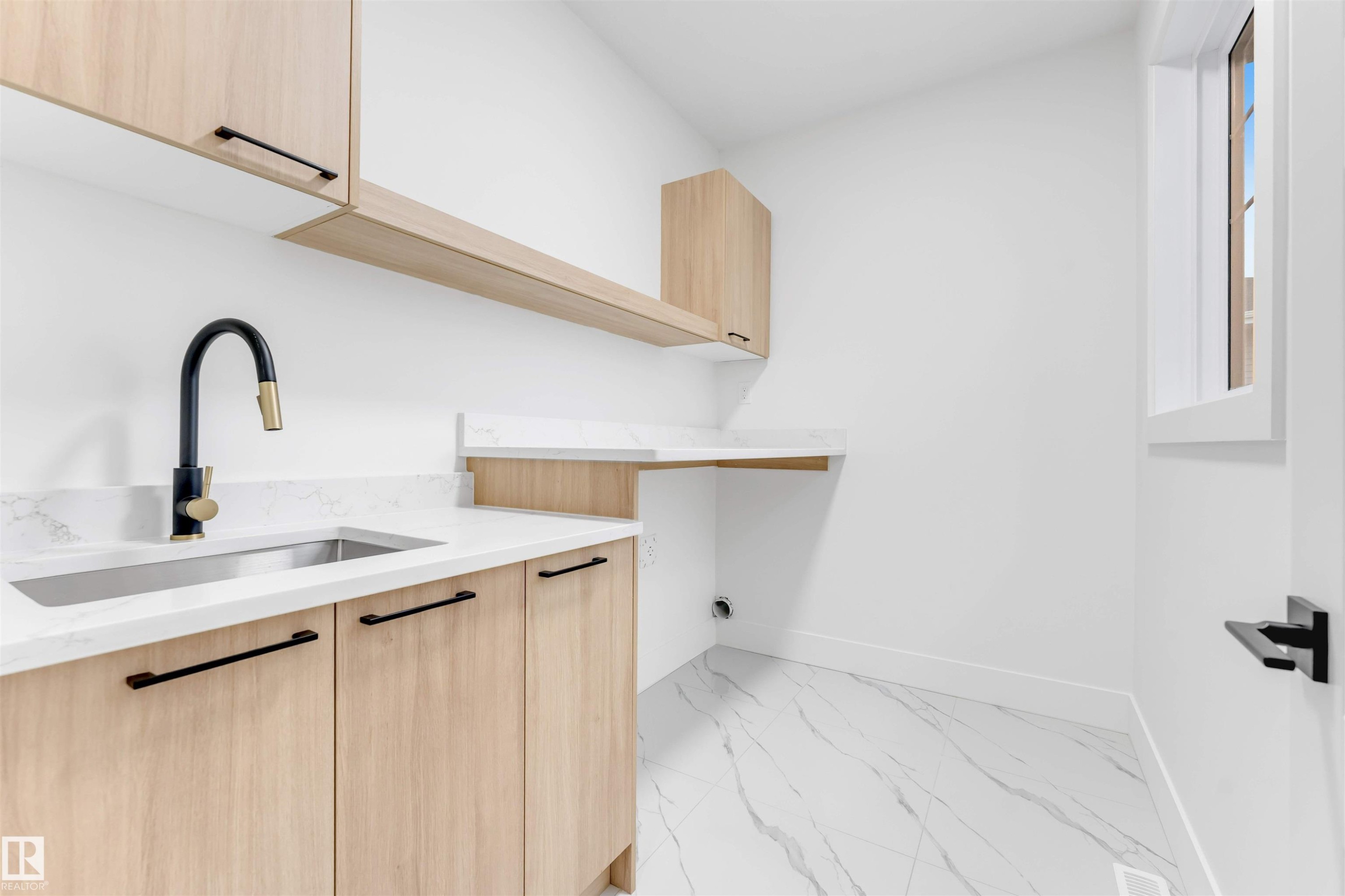 Well-appointed utility room featuring light wood cabinetry with black hardware, a stainless steel sink with a black and brass faucet, white countertops with veining, and white tiled flooring - 4004 35 Street, Beaumont, AB - Indoor Photo Showing Kitchen