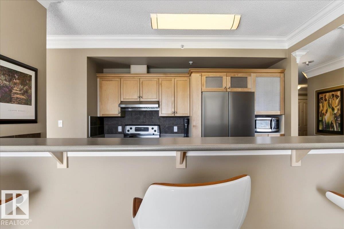 Kitchen featuring light wood cabinetry, stainless steel appliances, dark tile backsplash, a range hood, and a breakfast bar with an overhang - 701 9707 105 Street, Edmonton, AB - Indoor Photo Showing Kitchen