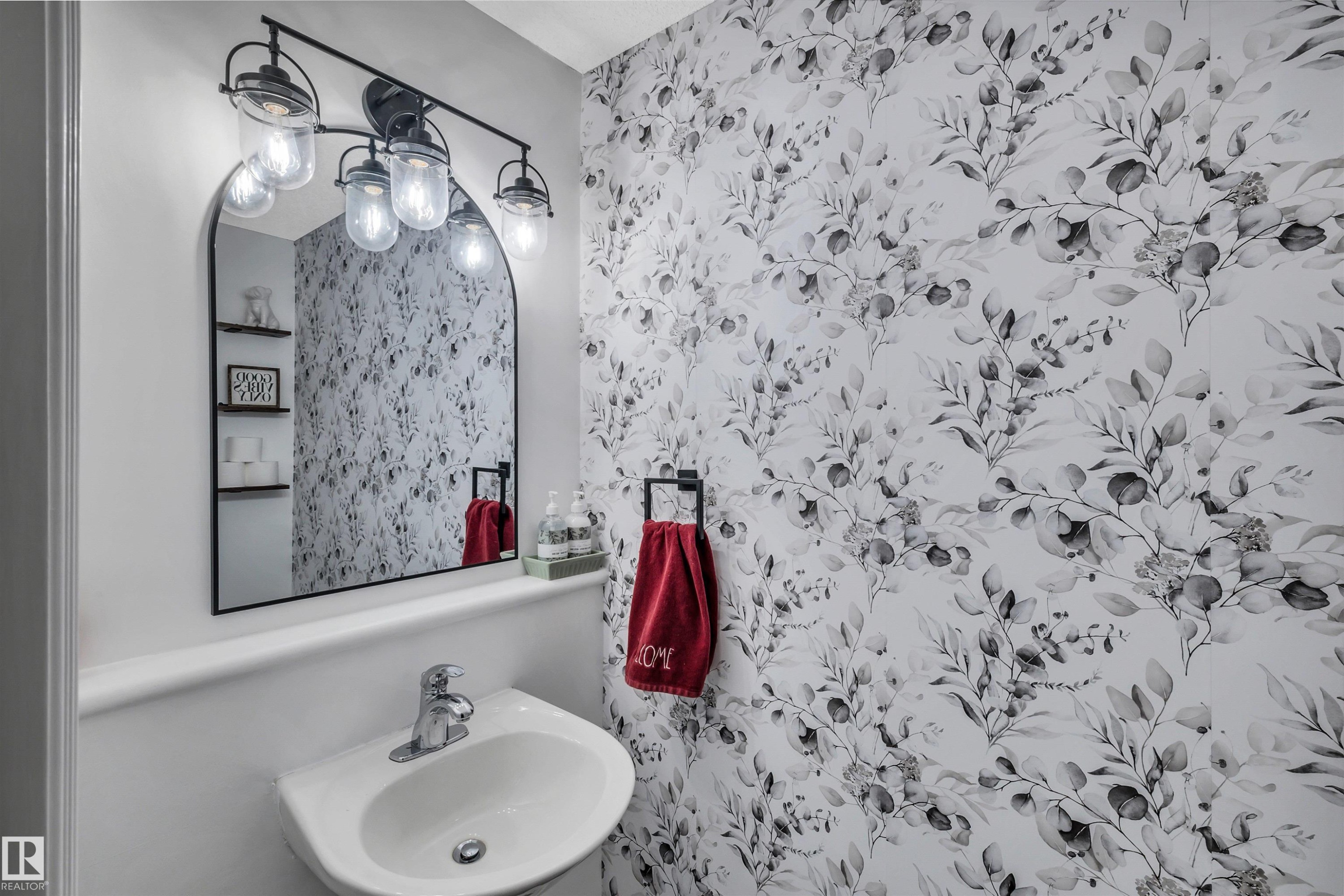 Powder room featuring botanical-patterned wallpaper, a wall-mounted sink with a chrome faucet, and an arched black-framed mirror - 96 Bonin Crescent, Beaumont, AB - Indoor Photo Showing Bathroom