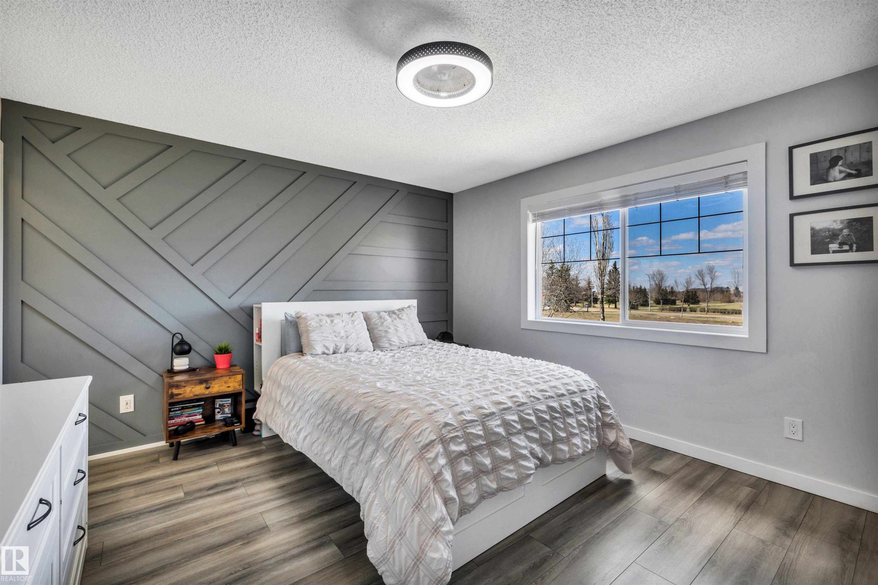 Feature wall with geometric paneling, wood-finish flooring, white trim, and a window framing outdoor scenery - 96 Bonin Crescent, Beaumont, AB - Indoor Photo Showing Bedroom