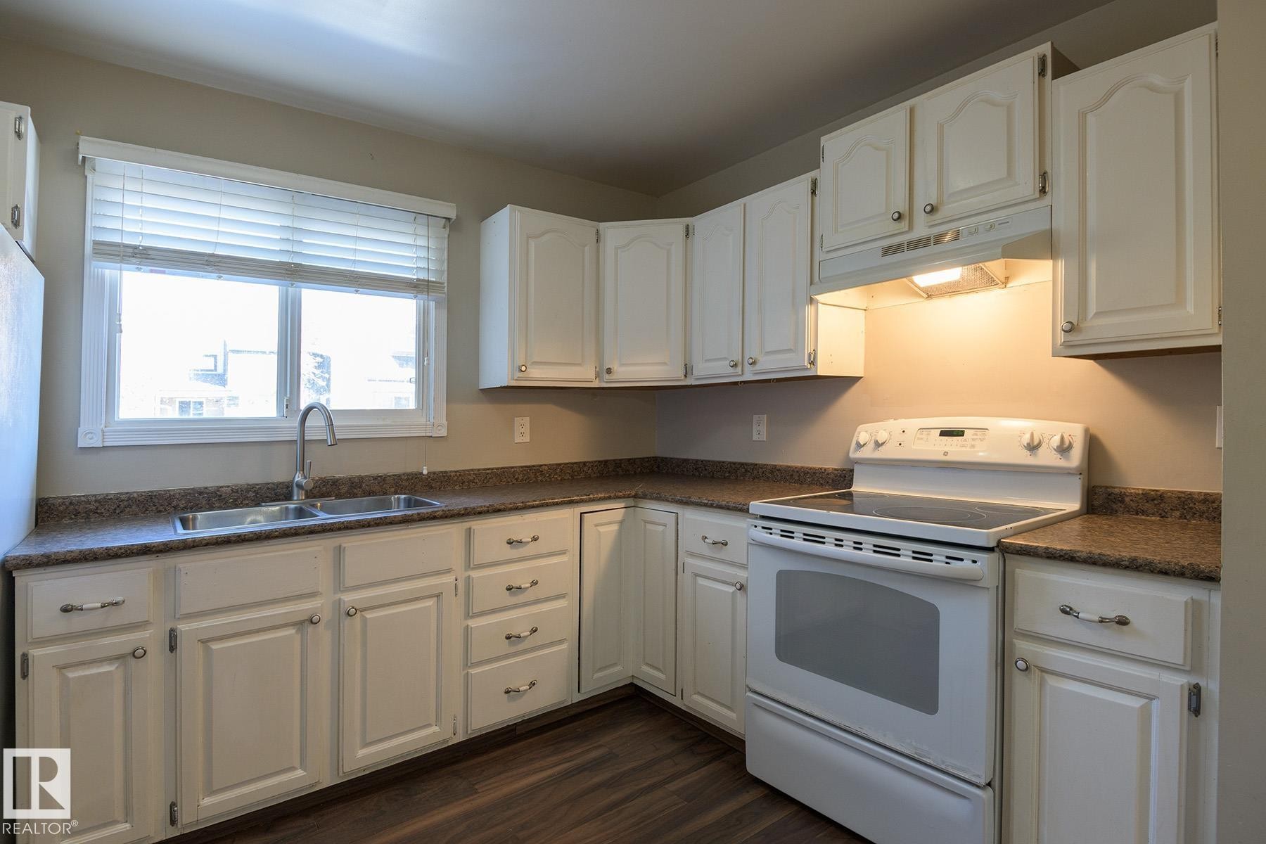 Kitchen featuring white cabinetry, ample counter space with a durable finish, a double basin stainless steel sink with a gooseneck faucet, a white electric range with an overhead microwave, and wood-finish flooring - 8039 27 Avenue, Edmonton, AB - Indoor Photo Showing Kitchen With Double Sink