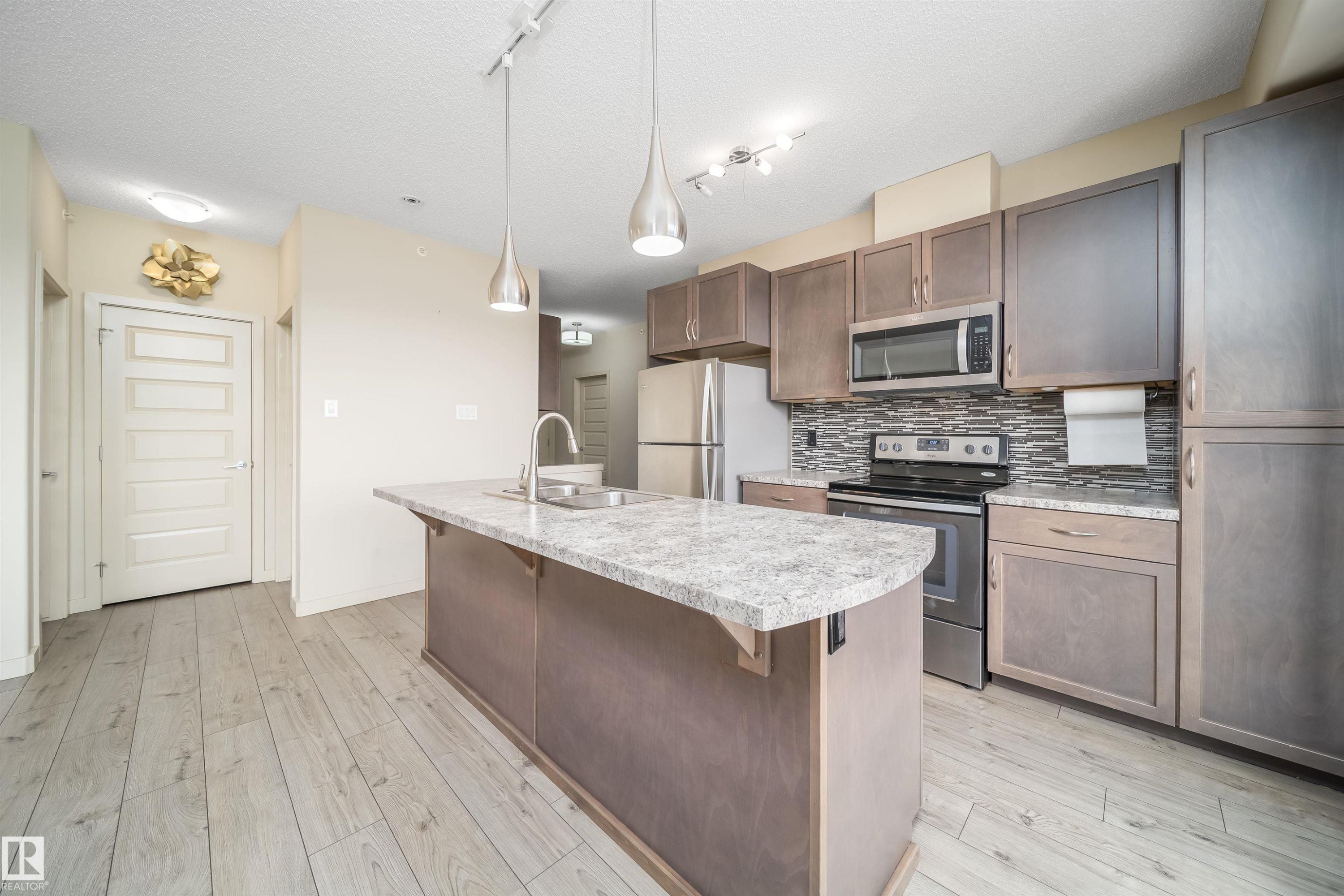 Open-concept kitchen featuring a central island with a granite-style countertop, stainless steel appliances, wood-finish cabinetry, a mosaic tile backsplash, and wood-finish flooring - 421 4008 Savaryn Drive, Edmonton, AB - Indoor Photo Showing Kitchen With Upgraded Kitchen