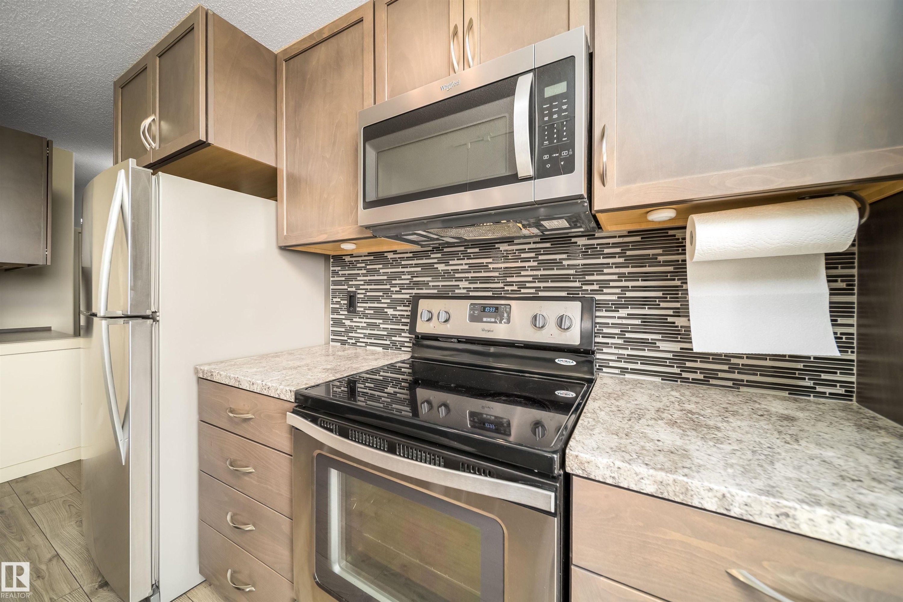 Kitchen featuring wood-finish cabinetry, speckled countertops, and a mosaic tile backsplash - 421 4008 Savaryn Drive, Edmonton, AB - Indoor Photo Showing Kitchen With Stainless Steel Kitchen With Upgraded Kitchen