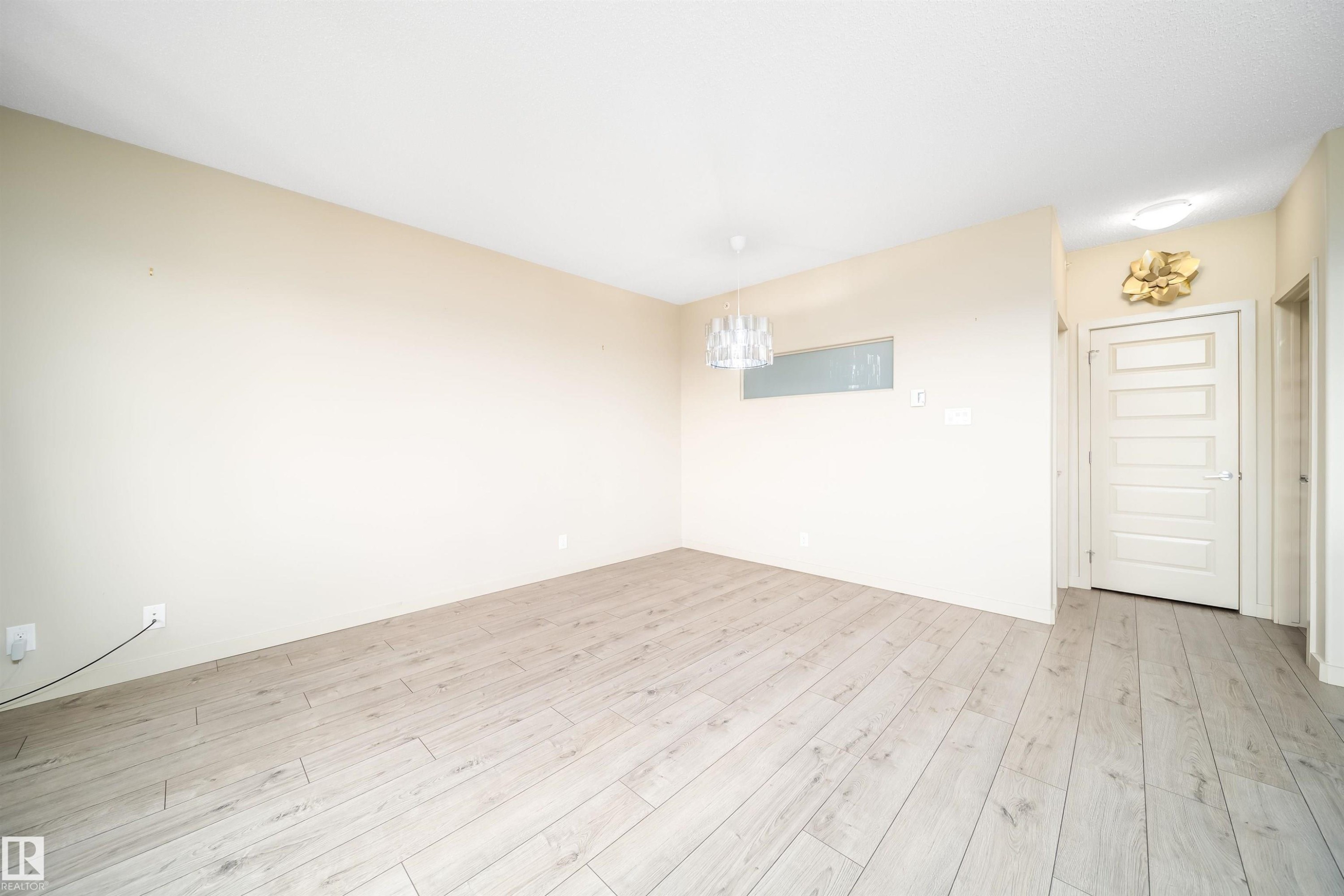 Spacious room featuring light wood-finish flooring, a contemporary chandelier, a white five-panel door, and neutral-toned walls - 421 4008 Savaryn Drive, Edmonton, AB - Indoor Photo Showing Other Room