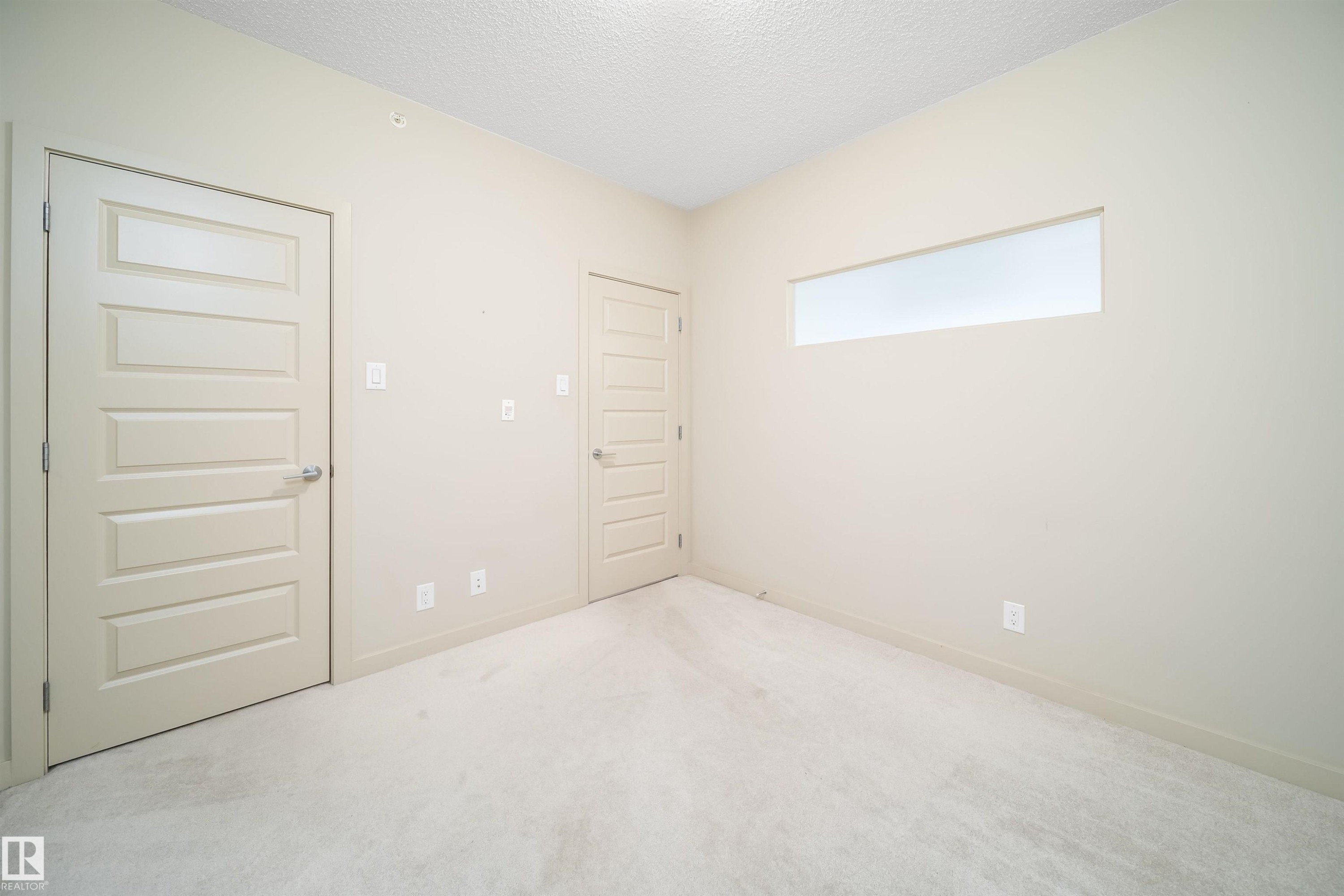 Neutral-toned room featuring a large rectangular transom window, light-colored carpet, and two five-panel interior doors with brushed nickel hardware - 421 4008 Savaryn Drive, Edmonton, AB - Indoor Photo Showing Other Room