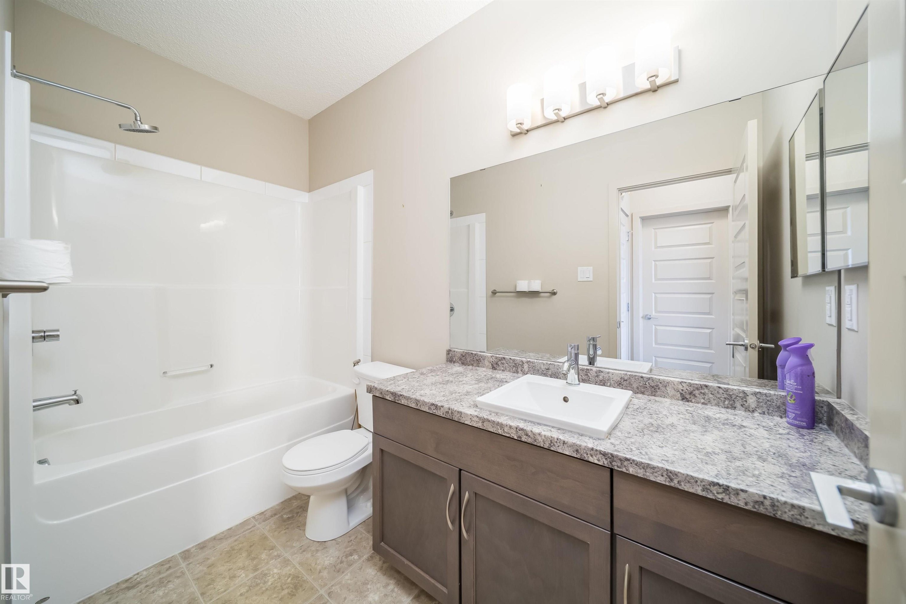 Bathroom featuring a white tub/shower combination, a toilet, and a vanity with a rectangular basin, light-colored patterned countertop, and brown cabinetry - 421 4008 Savaryn Drive, Edmonton, AB - Indoor Photo Showing Bathroom
