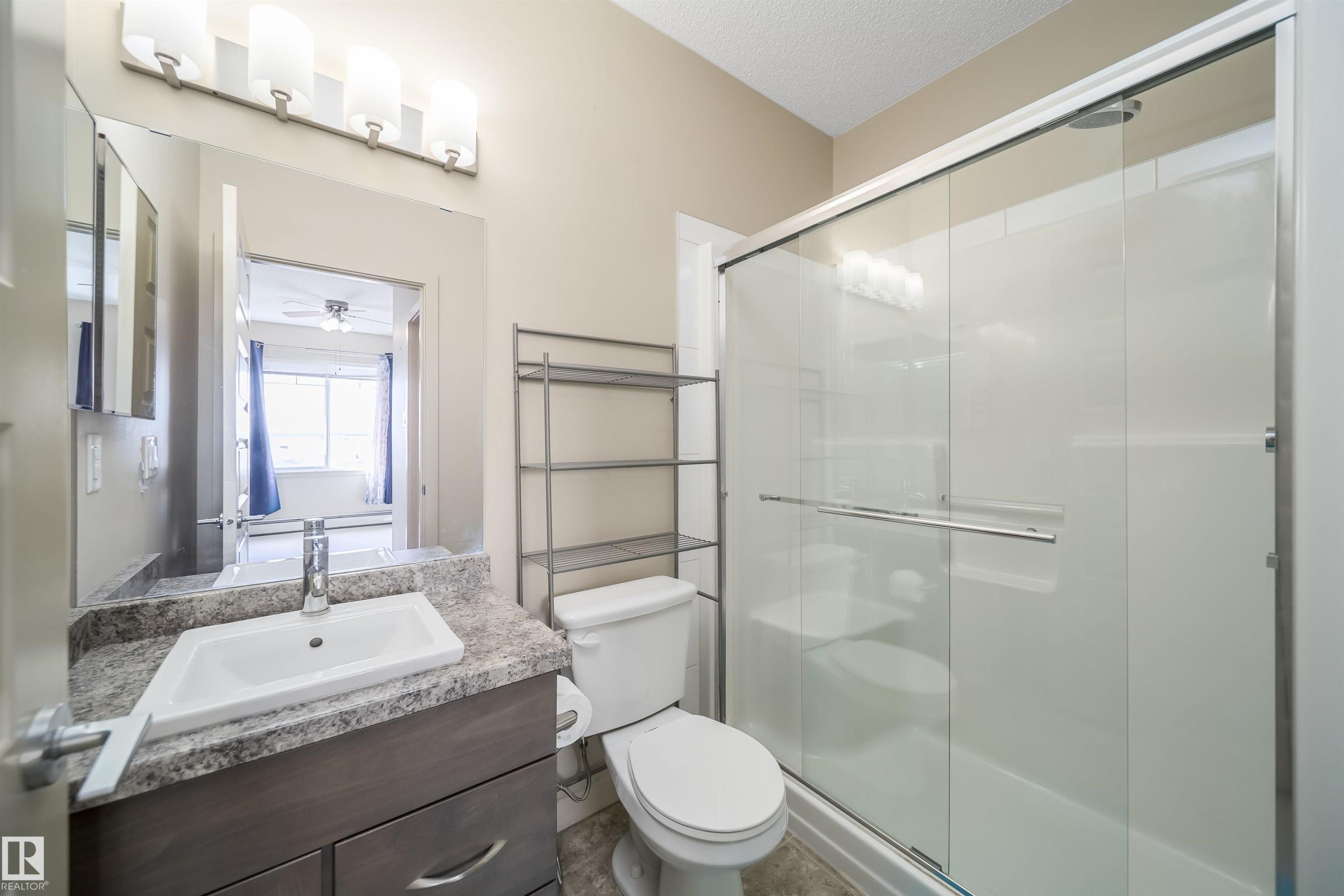 Bathroom featuring a sliding glass shower enclosure, a vanity with a rectangular basin sink, a speckled countertop, and a vanity light fixture with four sconces - 421 4008 Savaryn Drive, Edmonton, AB - Indoor Photo Showing Bathroom