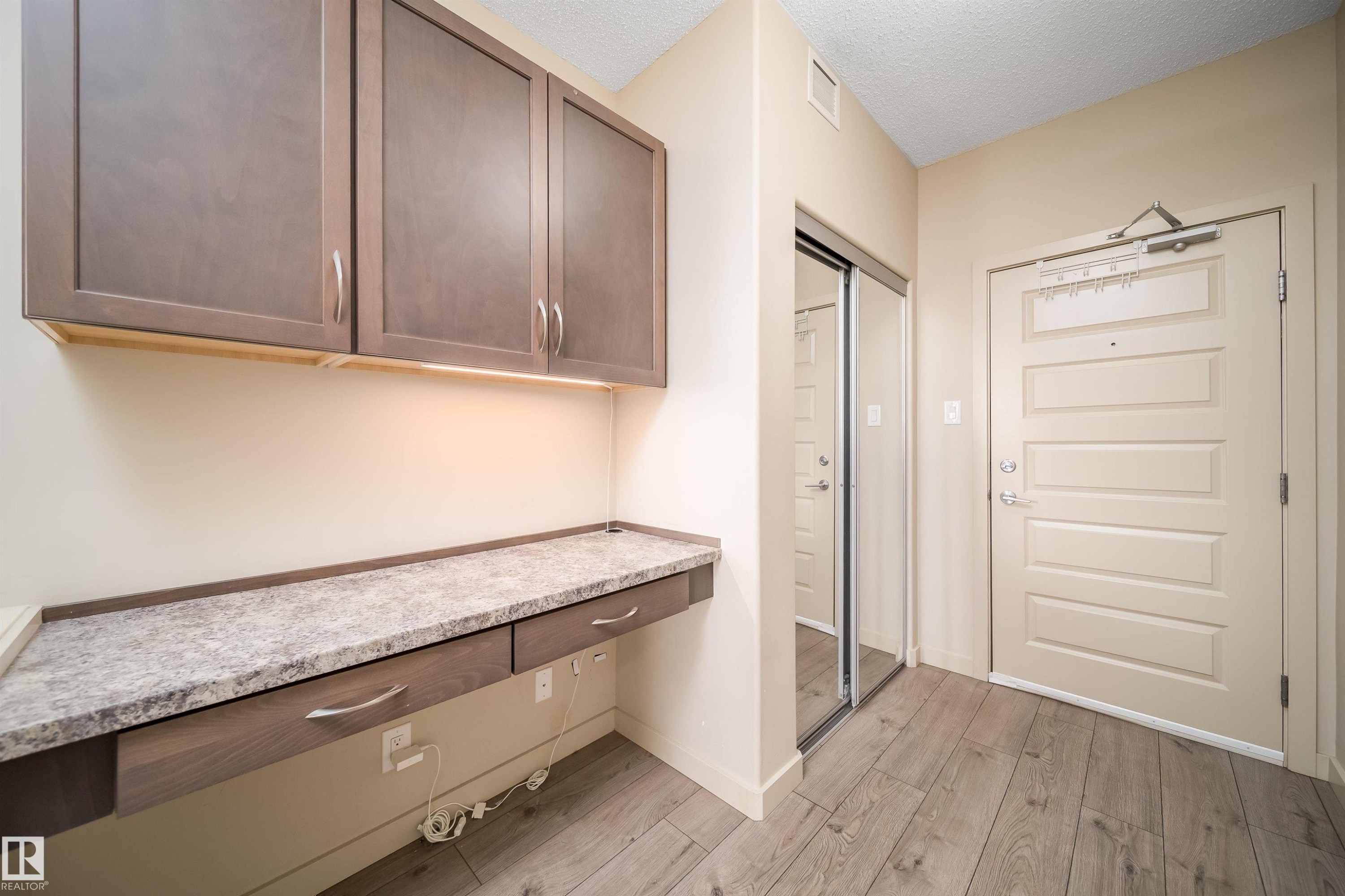 Entryway featuring wood-finish flooring, a built-in desk with two drawers and an integrated countertop, upper cabinetry with under-cabinet lighting, a mirrored sliding closet door, and a paneled entry door - 421 4008 Savaryn Drive, Edmonton, AB - Indoor Photo Showing Other Room