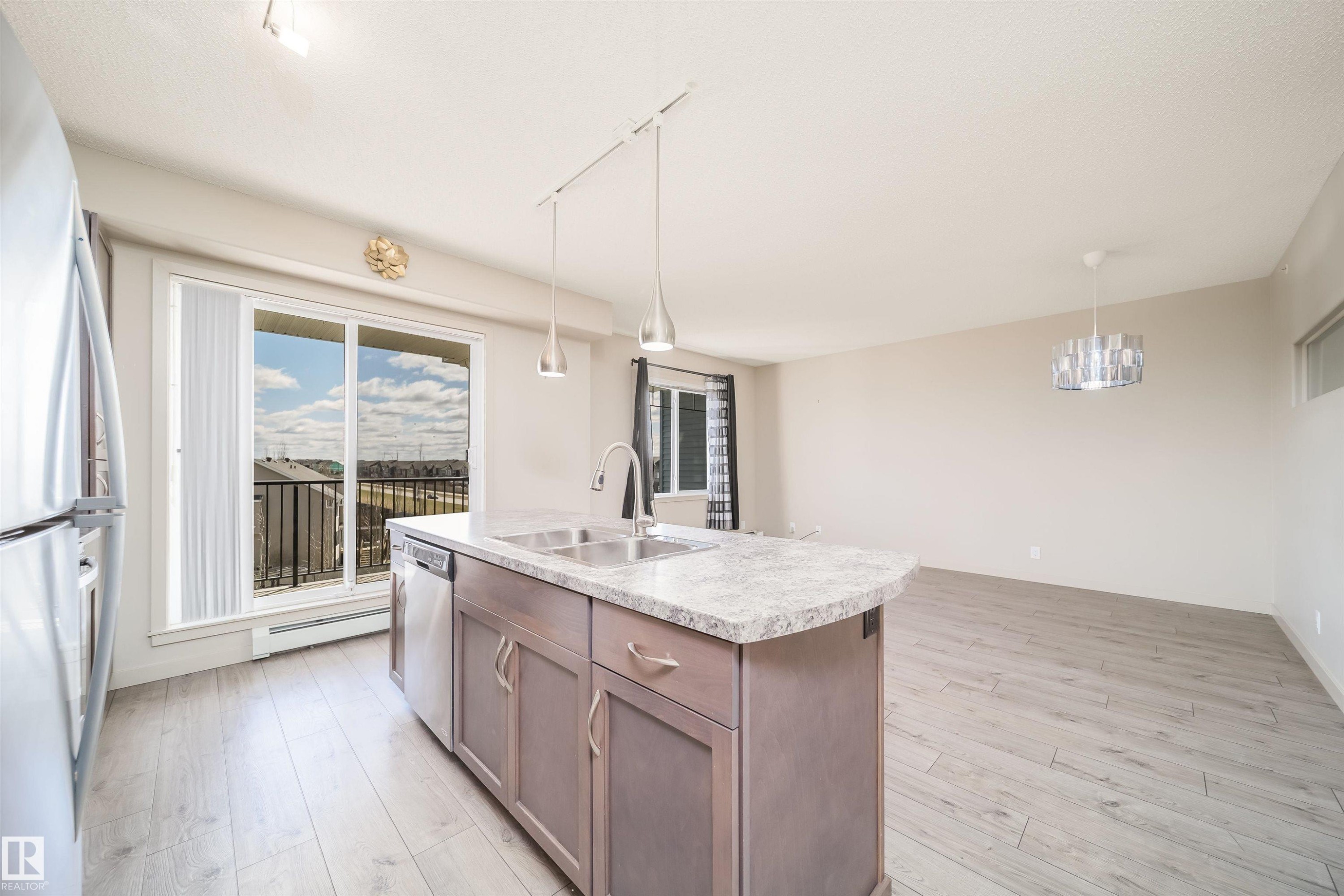 Open-concept living space featuring wood-finish flooring, a kitchen island with stone-finish countertop, stainless steel appliances, a double basin sink with a gooseneck faucet, and a balcony with black railings - 421 4008 Savaryn Drive, Edmonton, AB - Indoor Photo Showing Kitchen With Double Sink