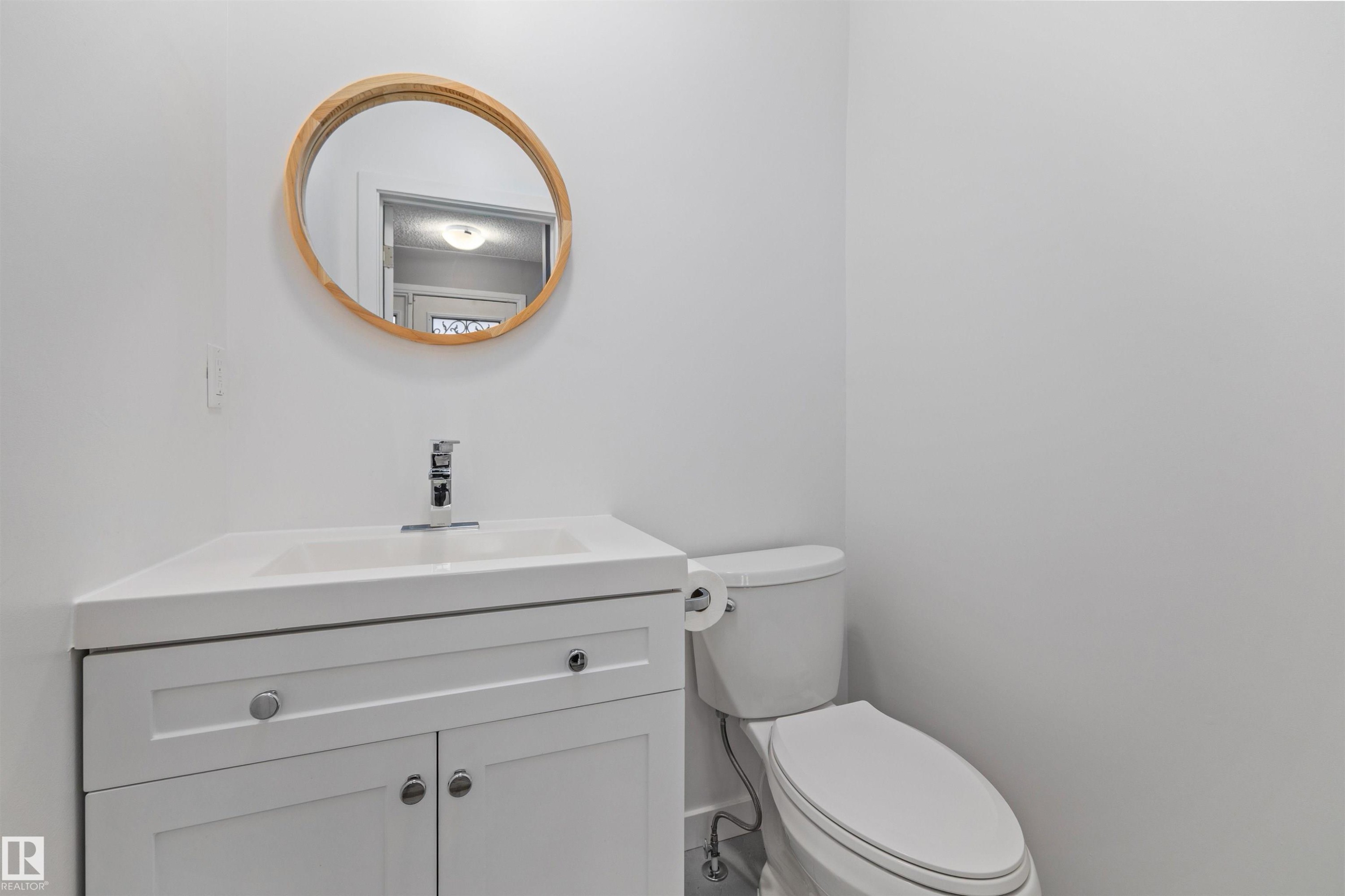 Powder room featuring a white single-sink vanity with a chrome faucet, white cabinetry with silver hardware, and a round wooden-framed mirror - 7927 71 Avenue, Edmonton, AB - Indoor Photo Showing Bathroom