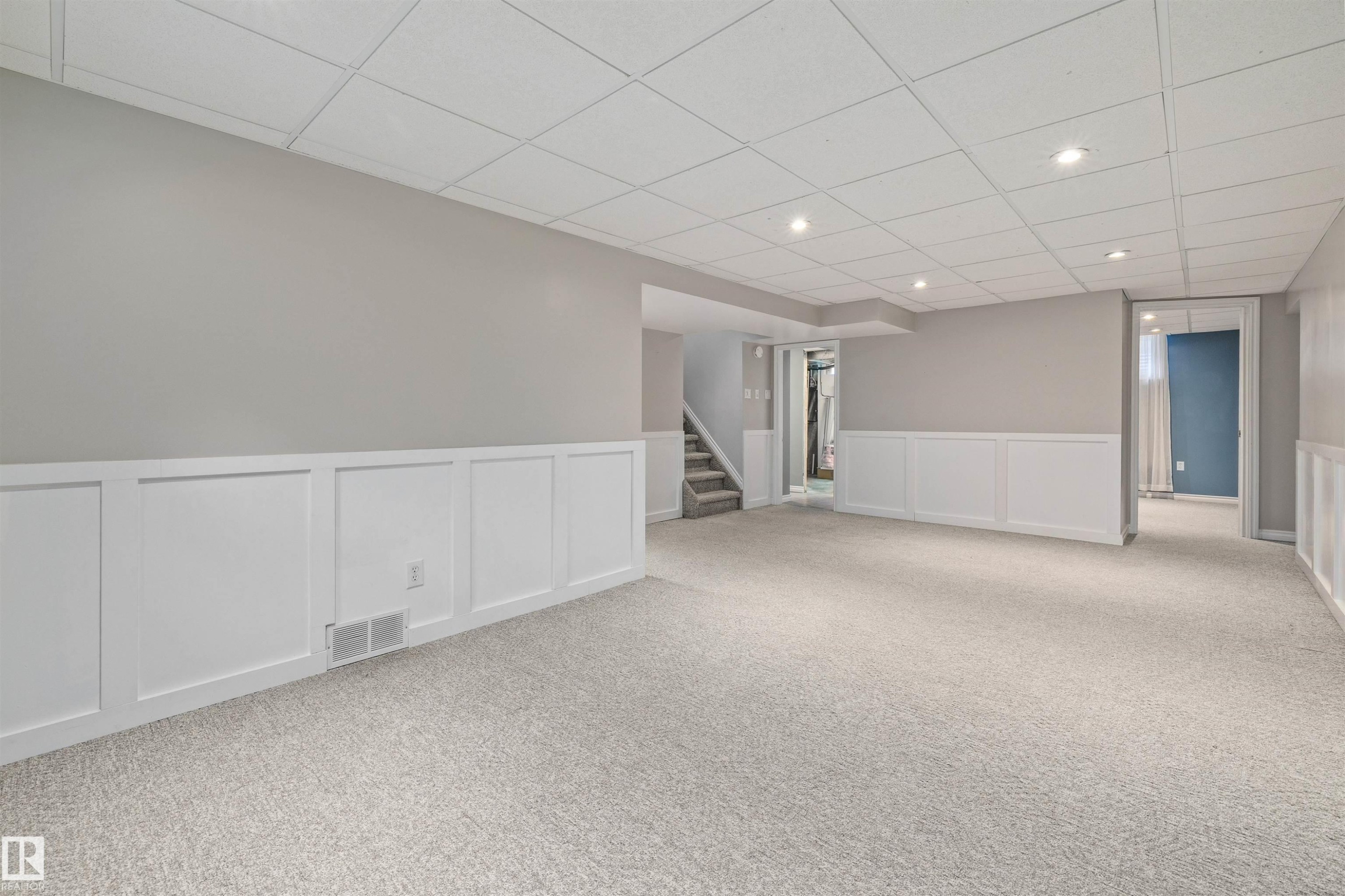 Spacious room featuring light gray carpet, white wainscoting, light gray walls, a white drop ceiling with recessed lighting, and a carpeted staircase - 7927 71 Avenue, Edmonton, AB - Indoor Photo Showing Other Room