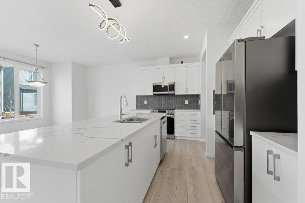 Spacious kitchen featuring an oversized island with a built-in sink, white cabinetry with contemporary hardware, stainless steel appliances, a dark tile backsplash, and light wood-finish flooring - 2011 207 Street, Edmonton, AB - Indoor Photo Showing Kitchen With Double Sink With Upgraded Kitchen