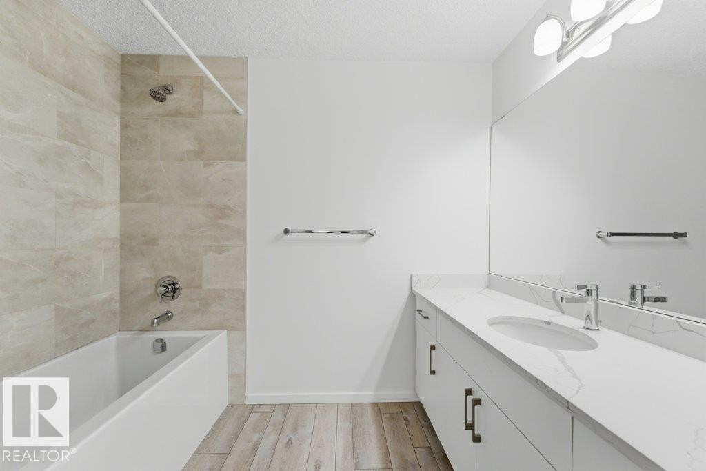 Bathroom featuring a white soaking tub with tiled surround, light-toned wood-finish flooring, a white vanity with an integrated sink, a full-width mirror, and brushed chrome fixtures - 2011 207 Street, Edmonton, AB - Indoor Photo Showing Bathroom