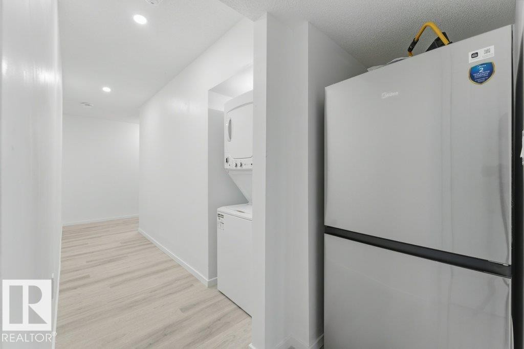 Bright white interior walls with recessed lighting, wood-finish flooring, a stainless steel refrigerator, and a stacked white washer and dryer unit - 2011 207 Street, Edmonton, AB - Indoor Photo Showing Laundry Room