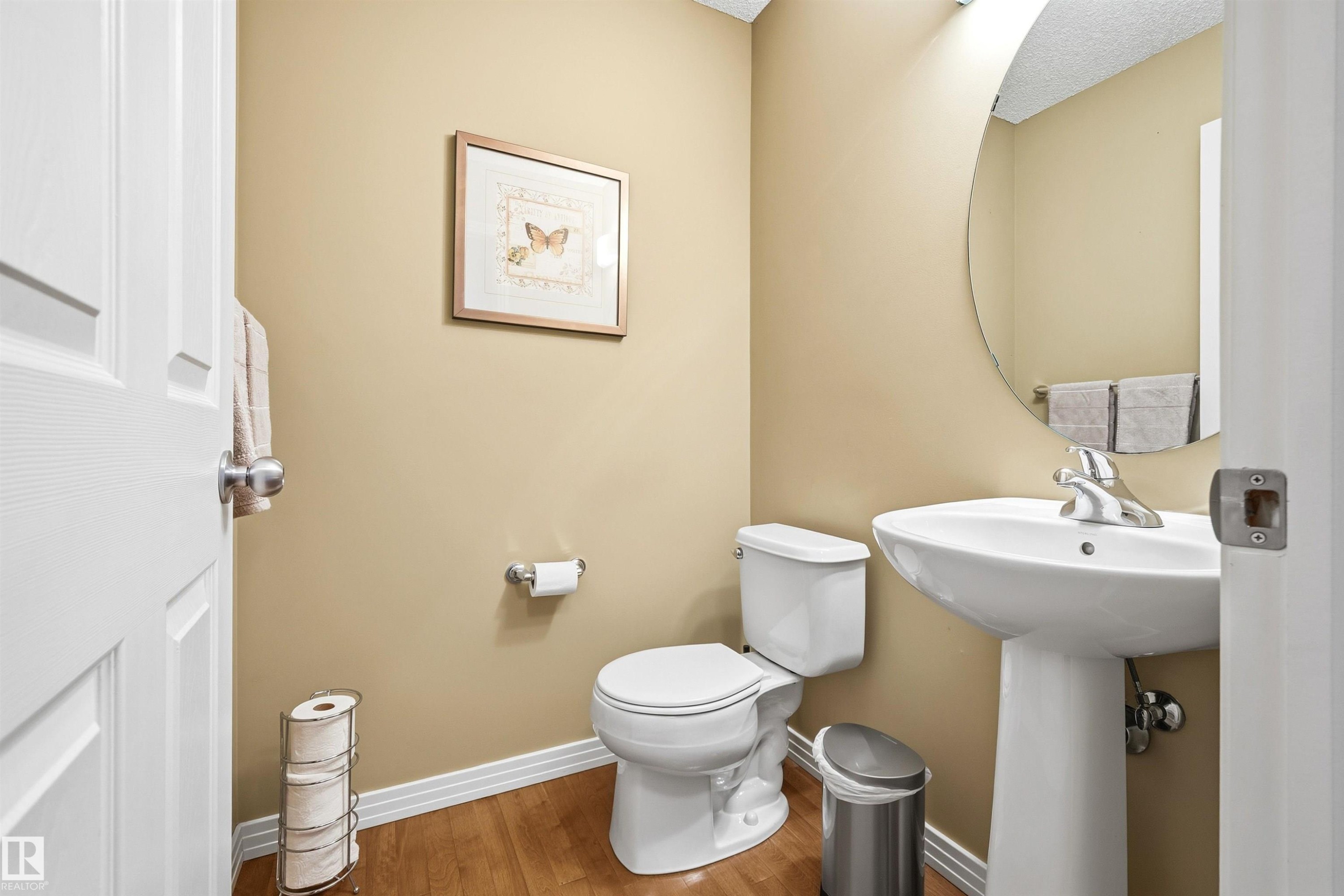 Pedestal sink with chrome faucet, circular mirror, and wood-finish flooring - 2340 Hagen Link, Edmonton, AB - Indoor Photo Showing Bathroom