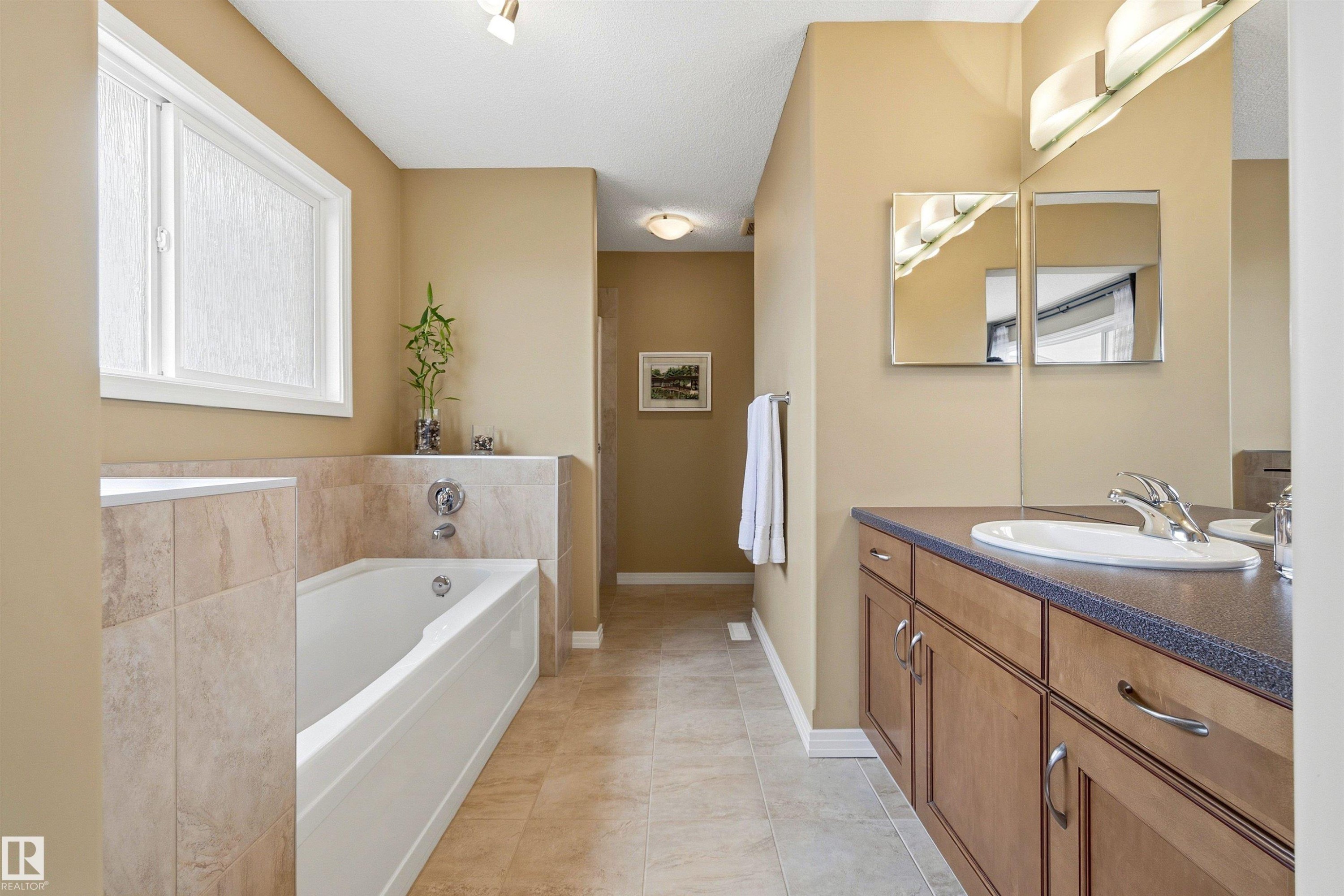 Bathroom featuring a built-in tub with tile surround, a window with obscure glass, and a vanity with wood-finish cabinetry, a white basin, and a wall-mounted mirror - 2340 Hagen Link, Edmonton, AB - Indoor Photo Showing Bathroom