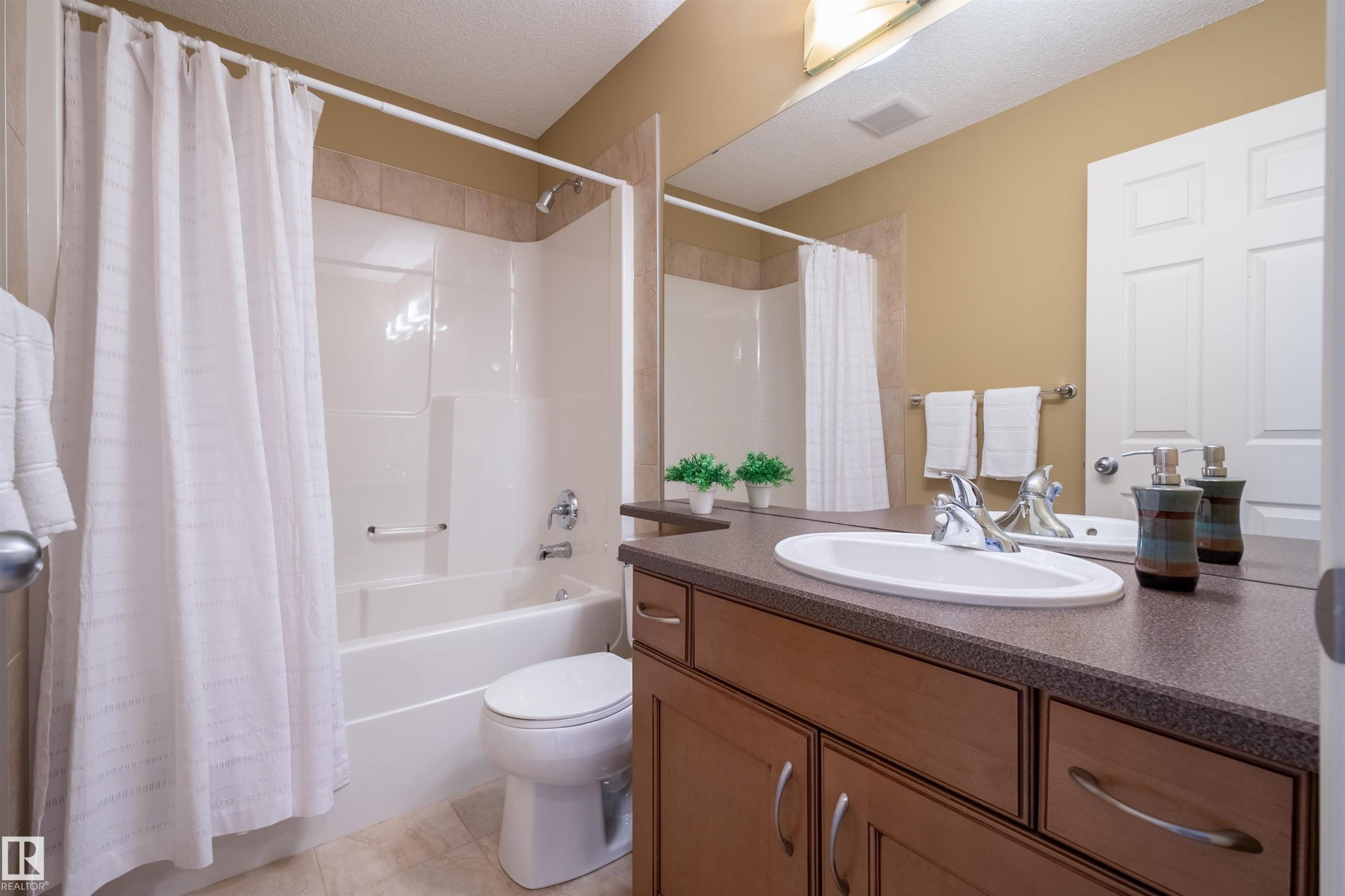 Bathroom featuring a white bathtub and shower combination, a single vanity with an oval sink, dark countertop, and wood-finish cabinetry - 2340 Hagen Link, Edmonton, AB - Indoor Photo Showing Bathroom