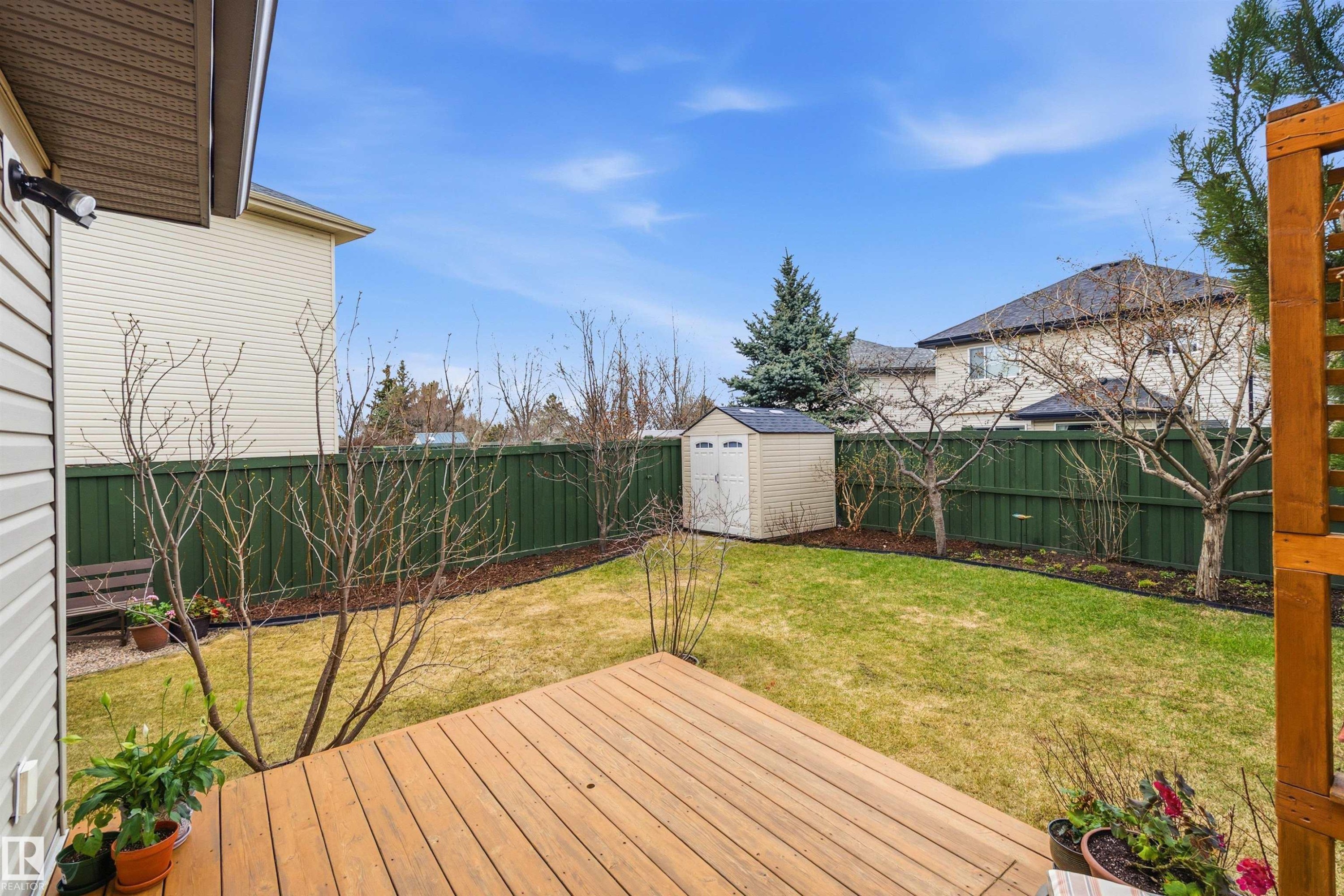 Expansive backyard featuring a wood-finish deck, green privacy fencing, mature trees, and a functional storage shed - 2340 Hagen Link, Edmonton, AB - Outdoor