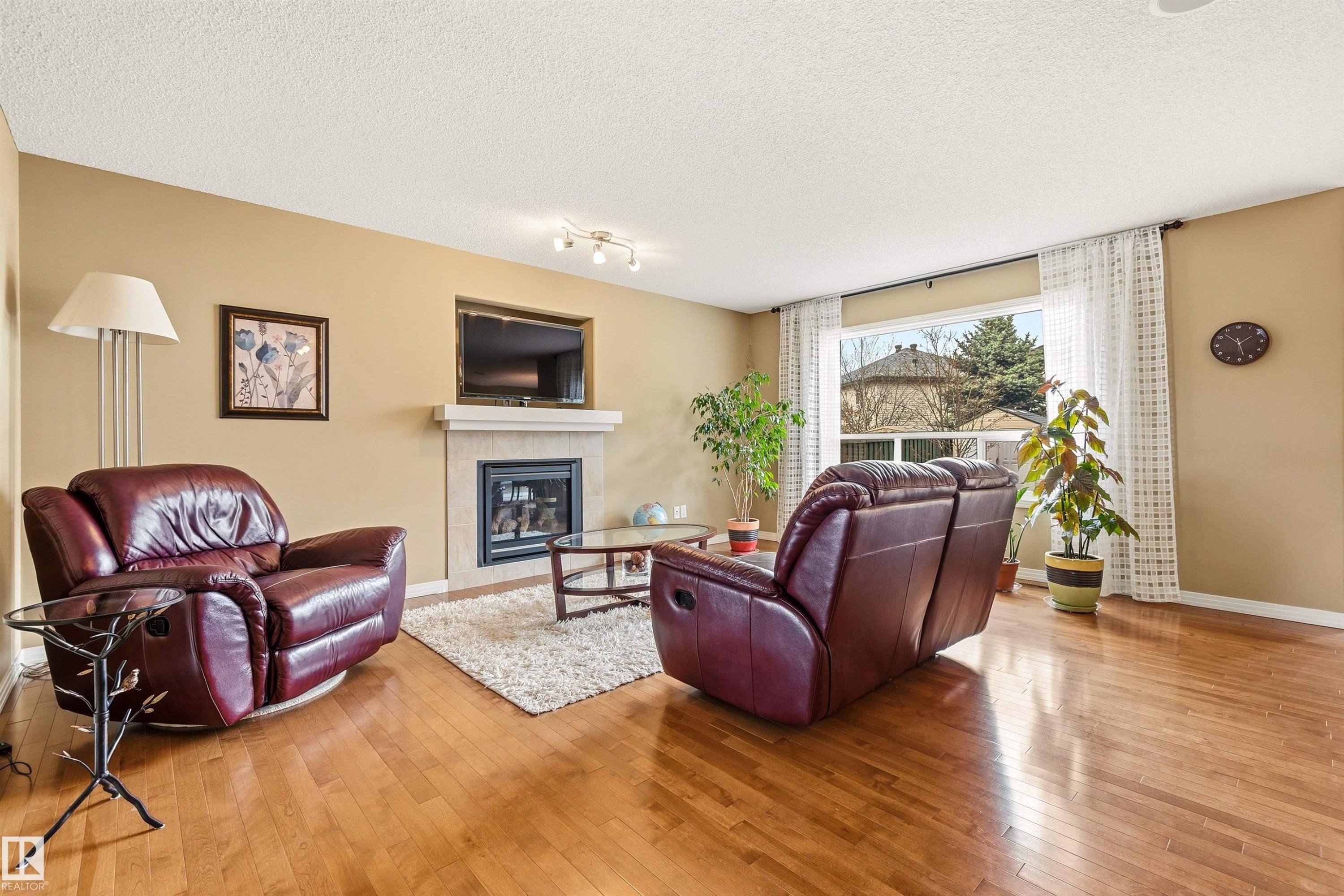 Living area featuring wood-finish flooring, a contemporary fireplace with a stone surround and mantel, and a large window with white curtains - 2340 Hagen Link, Edmonton, AB - Indoor Photo Showing Living Room With Fireplace