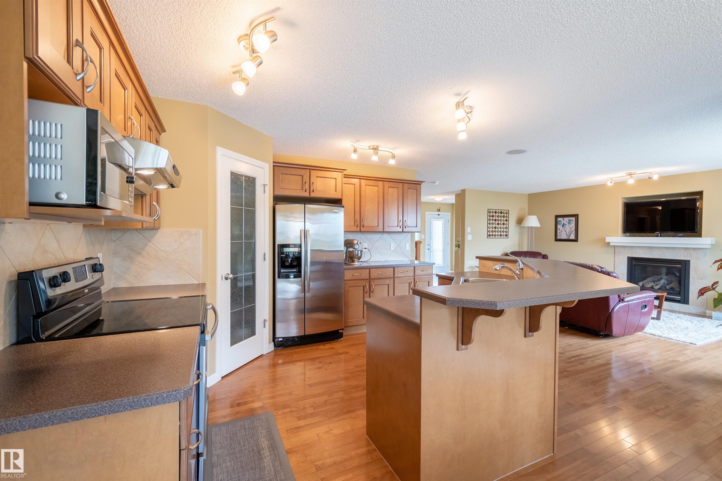 The kitchen features wood cabinetry, stainless steel appliances, a tile backsplash, and an island with an integrated sink - 2340 Hagen Link, Edmonton, AB - Indoor Photo Showing Kitchen