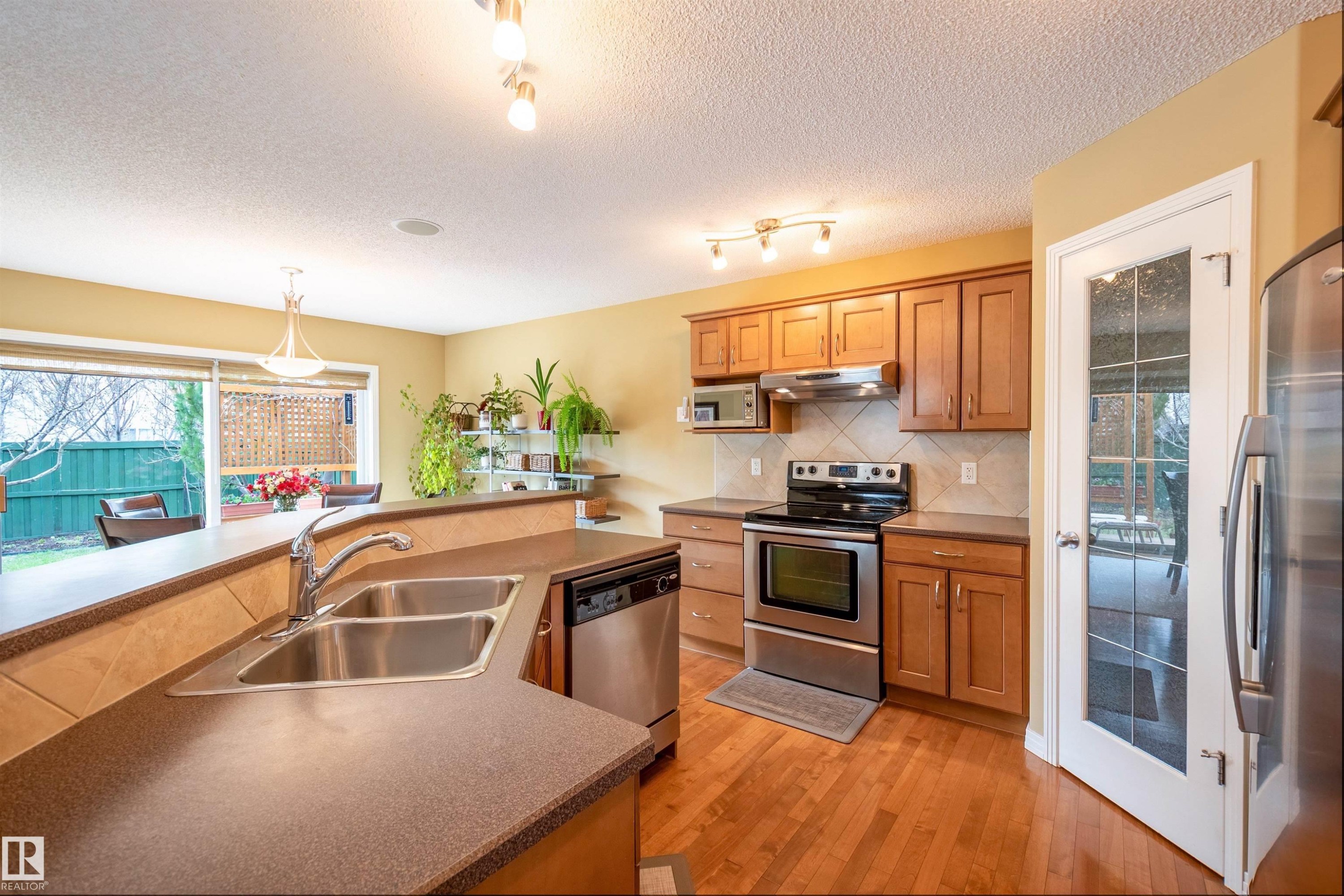 Kitchen featuring wood-finish flooring, stainless steel appliances, wood cabinetry, a double basin sink, and a glass-paned door - 2340 Hagen Link, Edmonton, AB - Indoor Photo Showing Kitchen With Double Sink