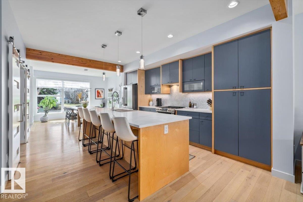 Open-concept kitchen featuring wood-finish flooring, a large island with a light-toned countertop, deep blue cabinetry with wood accents, pendant lighting, and a visible wood beam - 11245 64 Street, Edmonton, AB - Indoor Photo Showing Kitchen With Upgraded Kitchen