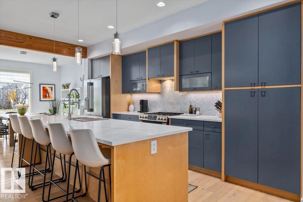 Modern kitchen featuring a large central island with a light-toned countertop, sleek dark cabinetry with wood-finish framing, under-cabinet lighting, a stainless steel range, and wood-finish flooring - 11245 64 Street, Edmonton, AB - Indoor Photo Showing Kitchen With Upgraded Kitchen
