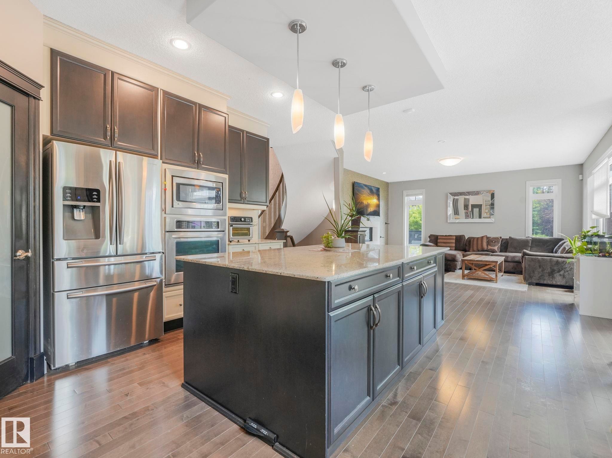 Gourmet kitchen featuring a large island with granite-style countertop, stainless steel appliances, dark wood-finish cabinetry, and recessed lighting, opening to a spacious living area with wood-finish flooring - 932 Summerside Link, Edmonton, AB - Indoor Photo Showing Kitchen With Upgraded Kitchen