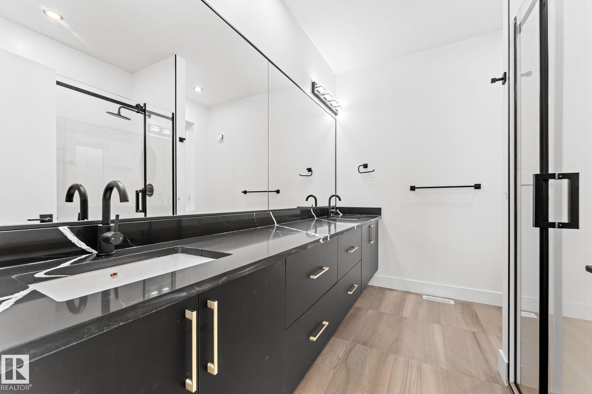 Modern bathroom featuring a double vanity with a dark countertop and gold hardware, a large mirror, and a glass-enclosed shower with black framing - 11019 132 St, Edmonton, AB - Indoor Photo Showing Bathroom