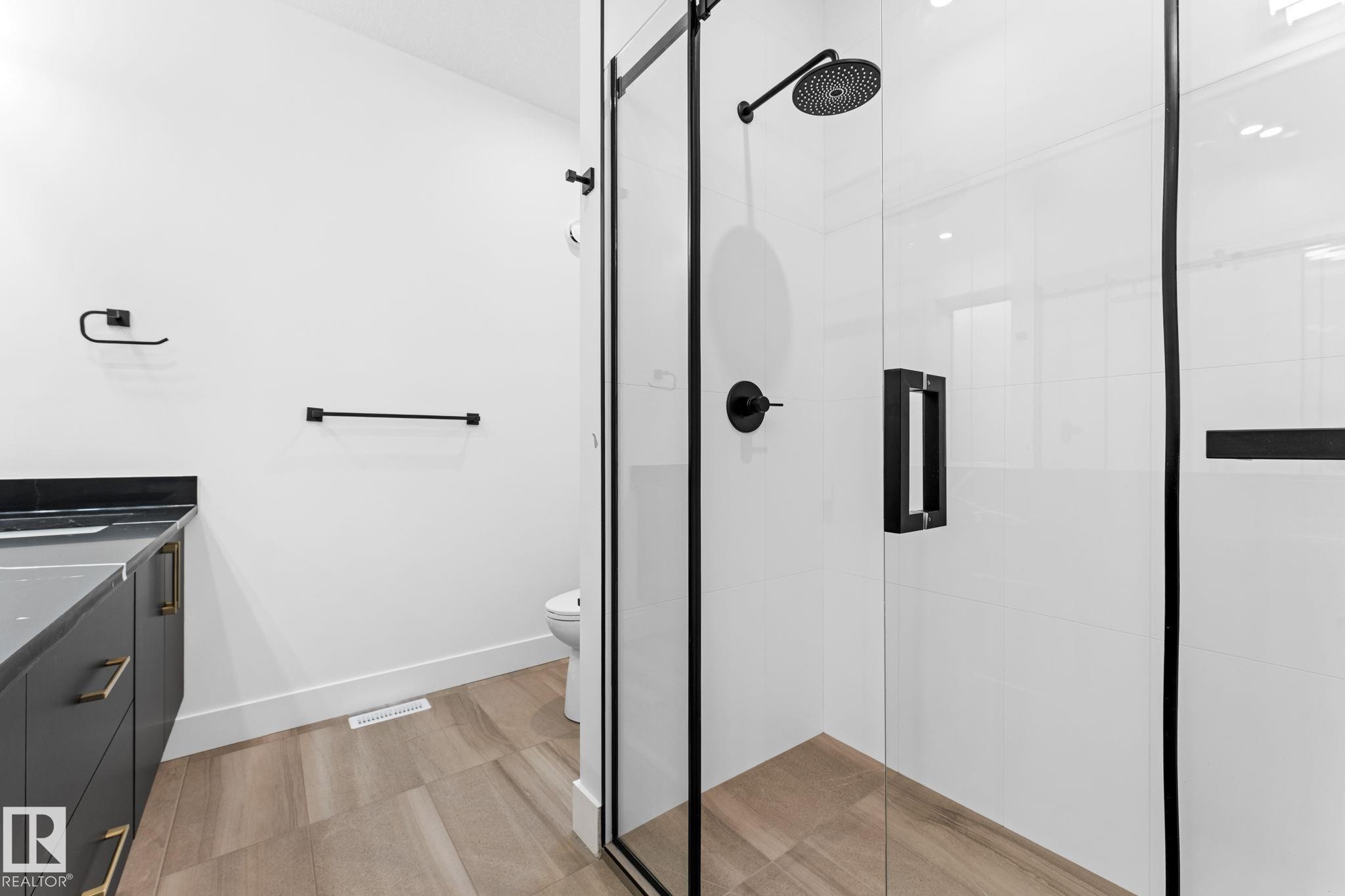 Modern bathroom featuring a glass-enclosed shower with matte black fixtures, a dark vanity with gold-toned handles and a stone countertop, and wood-look flooring - 11019 132 St, Edmonton, AB - Indoor