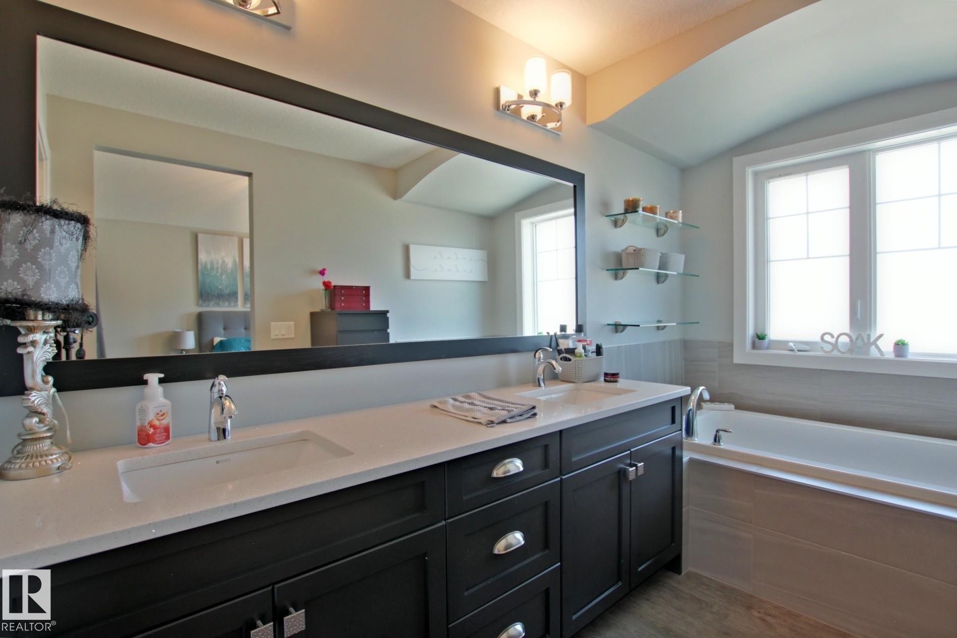 Bathroom featuring a dual vanity with a white countertop and dark cabinetry, a large framed mirror, a built-in bathtub with tiled surround, and multiple glass display shelves - 9711 223 Street, Edmonton, AB
