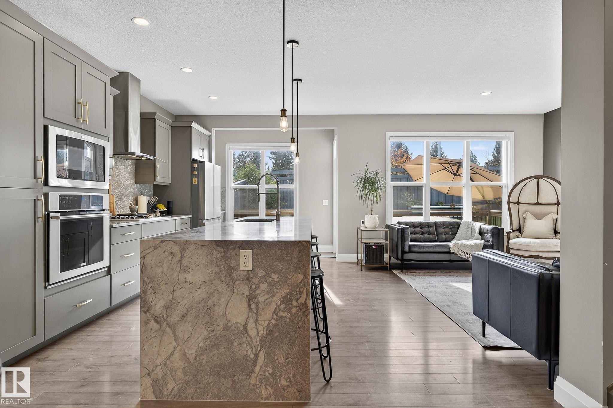 Open-concept kitchen and living area featuring a large central island with stone countertop, gray cabinetry, stainless steel appliances including a built-in oven and microwave, and wood-finish flooring - 1737 Dumont Crescent, Edmonton, AB - Indoor Photo Showing Kitchen With Upgraded Kitchen