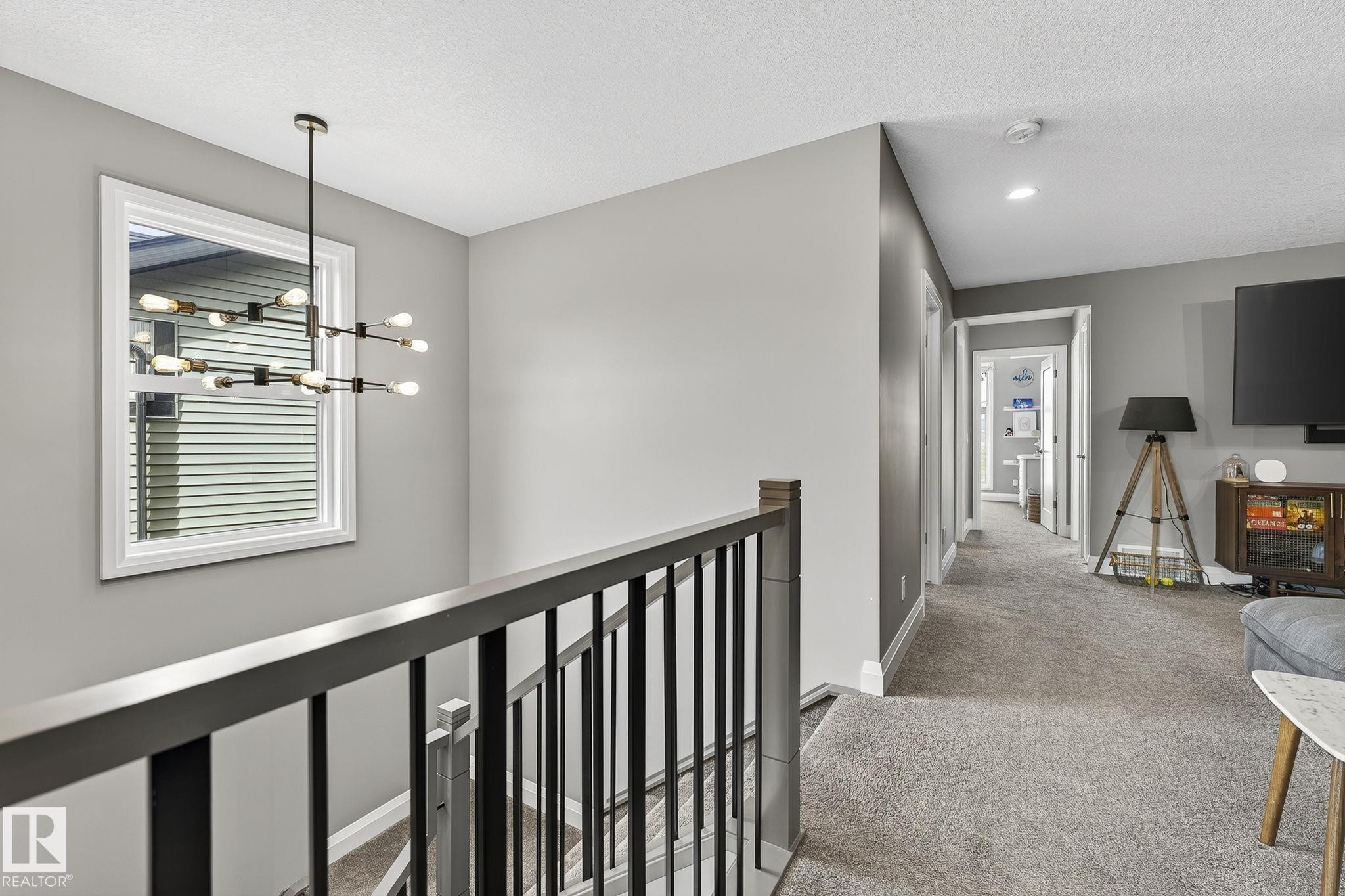 Upper-level landing featuring a contemporary chandelier, a window with white trim, dark wood-finish handrails with black balusters, plush gray carpeting, and recessed lighting - 1737 Dumont Crescent, Edmonton, AB - Indoor Photo Showing Other Room