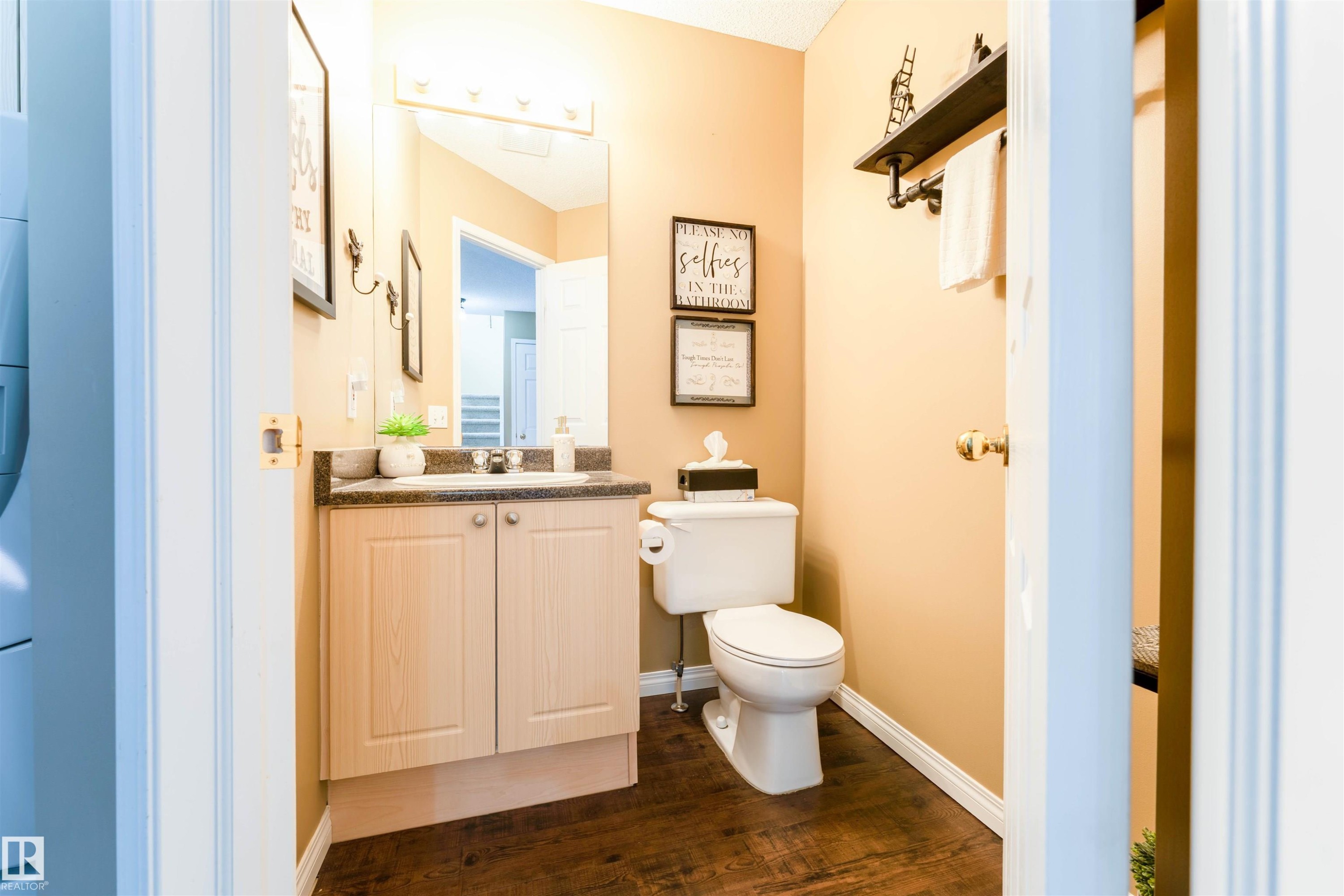 Powder room featuring a single vanity with a dark countertop and light wood-finish cabinetry, a mirror with overhead lighting, a toilet, and dark wood-finish flooring - 847 Blacklock Way, Edmonton, AB - Indoor Photo Showing Bathroom