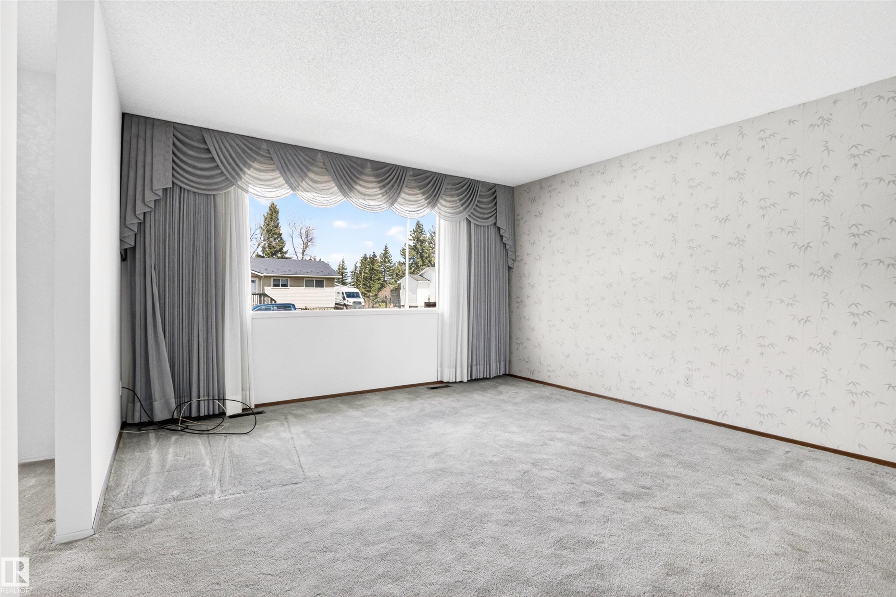 Spacious room featuring a large window with draped valance, light-toned patterned wallpaper, light gray carpeting, and white trim - 29 Bristow Crescent, Spruce Grove, AB - Indoor Photo Showing Other Room